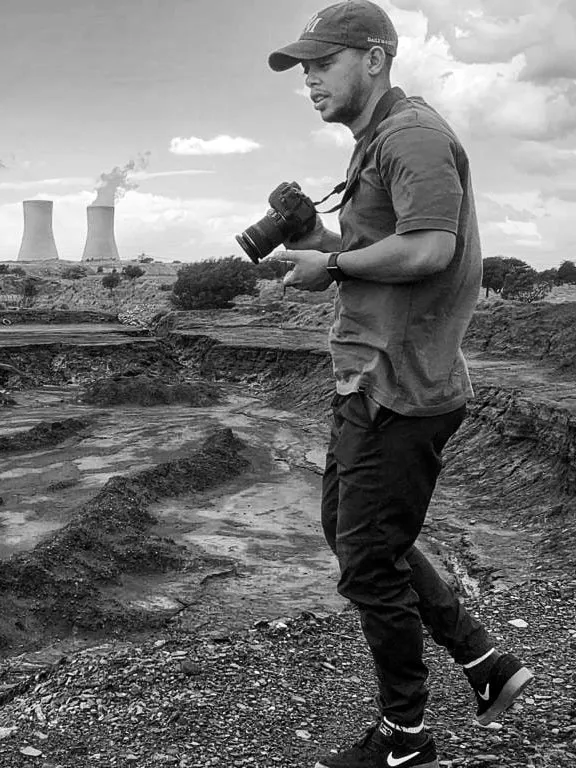 A man holds a camera in a field. There are smoke stacks in the background