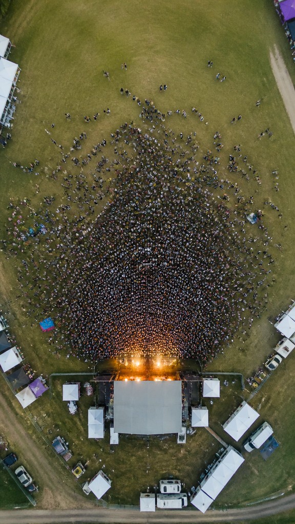 a live music scene with a glowing stage and silhouetted crowd, energetic vertical composition with open space at the top, no text — Aerial Drone Photography Style flyer