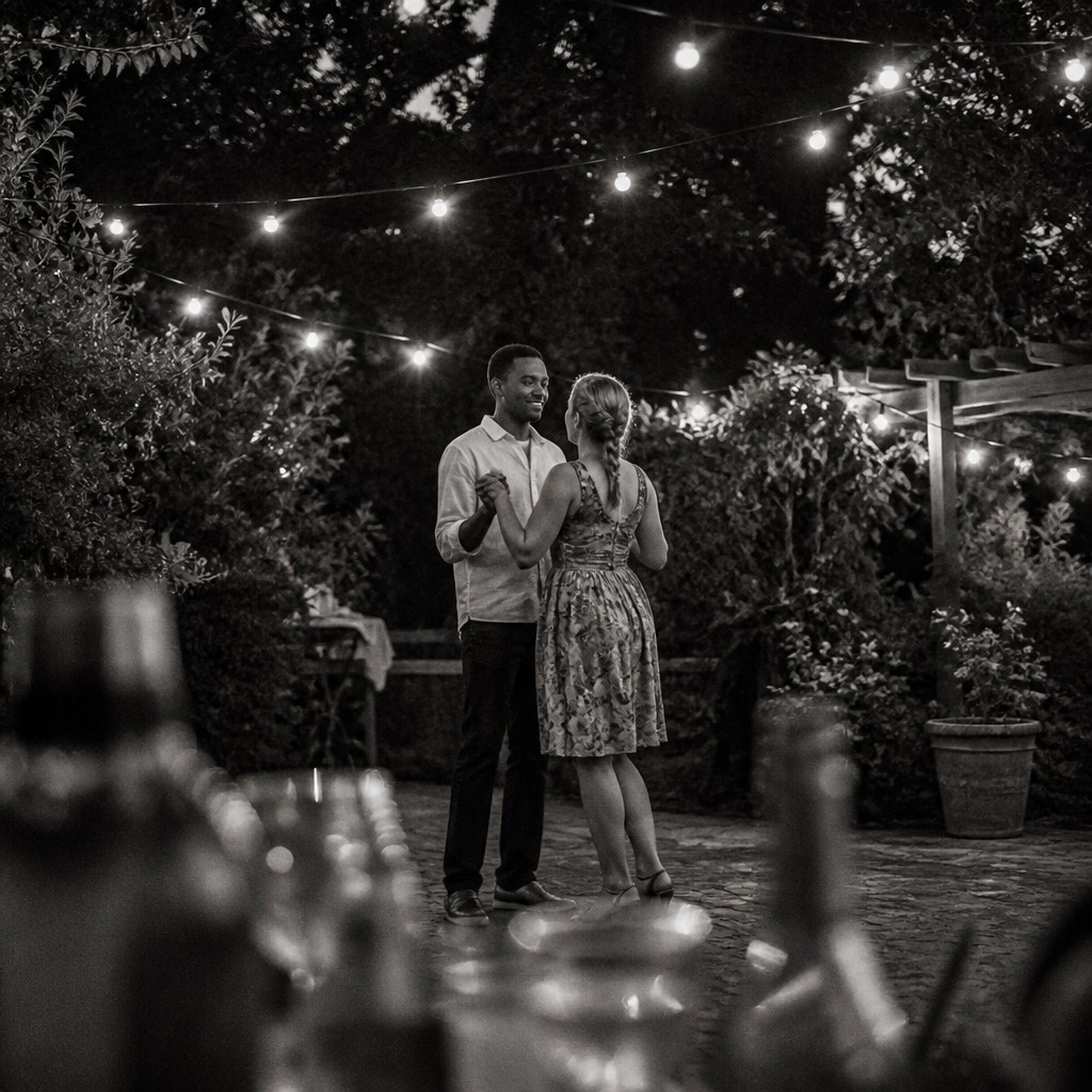 an original couple dancing under string lights in a garden at dusk: a man with short black hair in a linen shirt and a woman with braided red hair in a summer dress — Street Photography Style couple portrait
