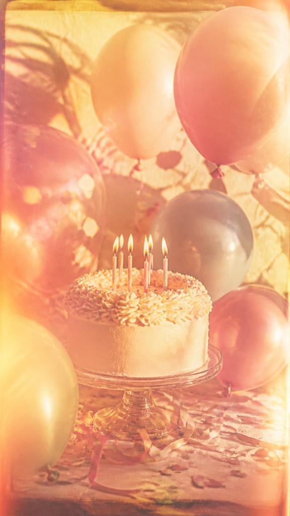 a birthday cake with lit candles surrounded by balloons and confetti, festive vertical composition, no text — Vintage Film Burn birthday card