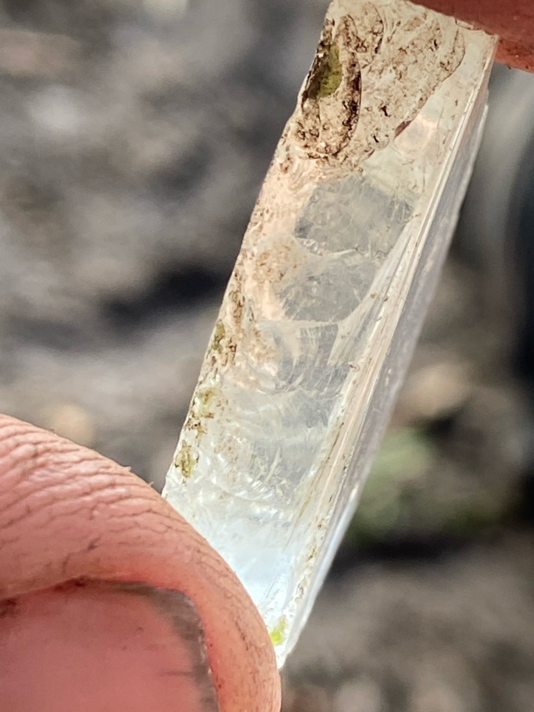 The image shows a raw mineral specimen, likely quartz, beryl, or a similar silicate crystal rather than a diamond. It displays conchoidal fractures, uneven surface textures, and mineral/dirt inclusions, which are inconsistent with natural diamond rough (octahedrons or dodecahedrons with curved faces). - Rough crystal / Irregular elongated fragment with flat cleavage or fractured planes