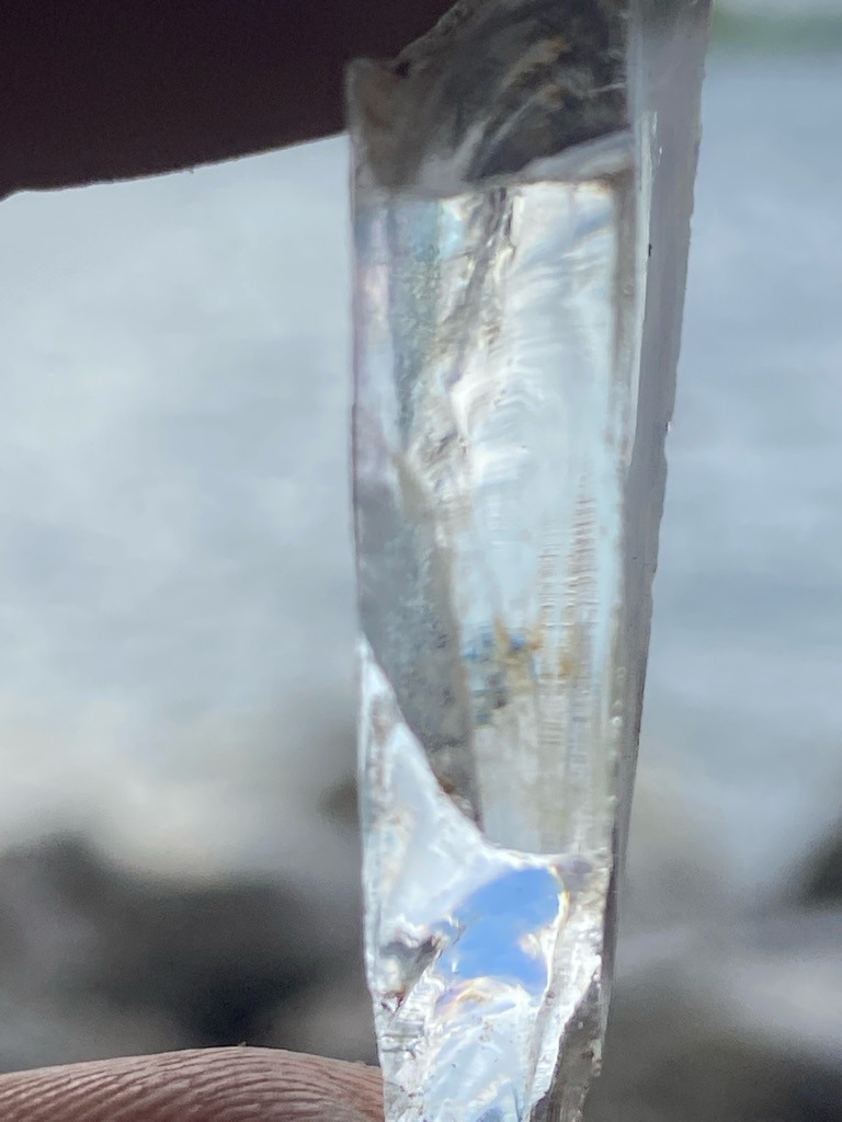 The specimen displayed in the image is not a diamond; it appears to be a raw piece of clear quartz crystal, glass, or potentially colorless beryl/topaz due to its elongated prismatic habit, striations on the crystal face, and conchoidal fracture pattern. - Raw, uncut crystal specimen with a naturally elongated, prismatic habit rather than a faceted gem shape.