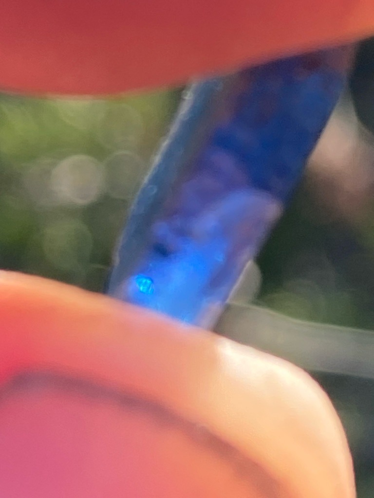 Natural Diamond (likely type IIa based on strong blue fluorescence under UV light, though visual assessment from a single blurry photo is limited) - Indeterminate / Rough or Raw Crystal (elongated crystal habit visible)