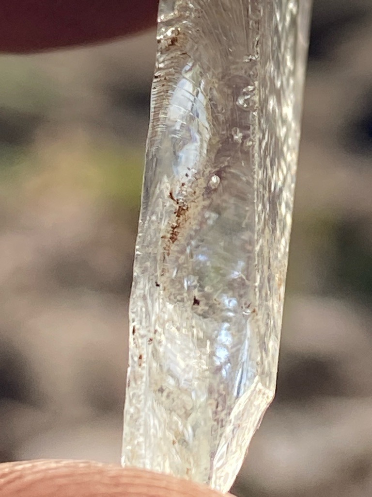 The image shows a raw mineral specimen, likely quartz or a similar crystalline rock, rather than a genuine faceted diamond. The striated growth lines and amorphous structure are inconsistent with rough diamond crystals. - Raw, unpolished elongated crystal fragment with irregular natural formation.