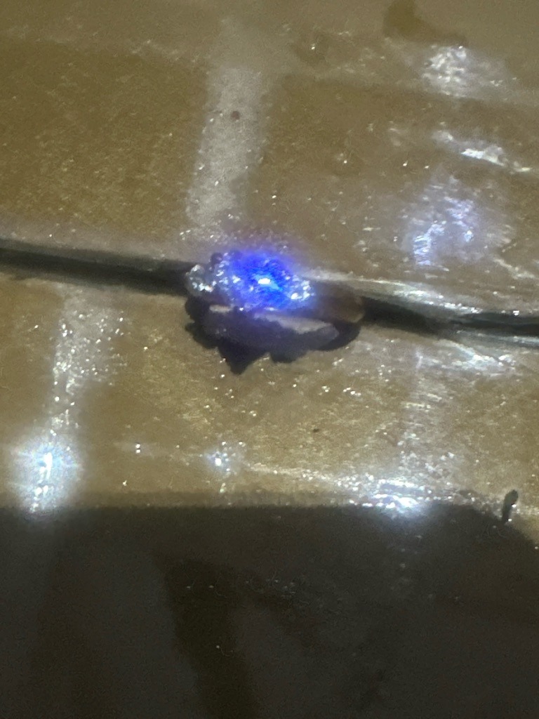 Unable to definitively determine diamond type from visual inspection under intense blue lighting; appears to be a mounted ring featuring a central stone with high refractive properties, potentially Moissanite or a treated/fluorescent diamond. - Round Brilliant (estimated from typical mounting styles, though exact faceting is obscured by glare)