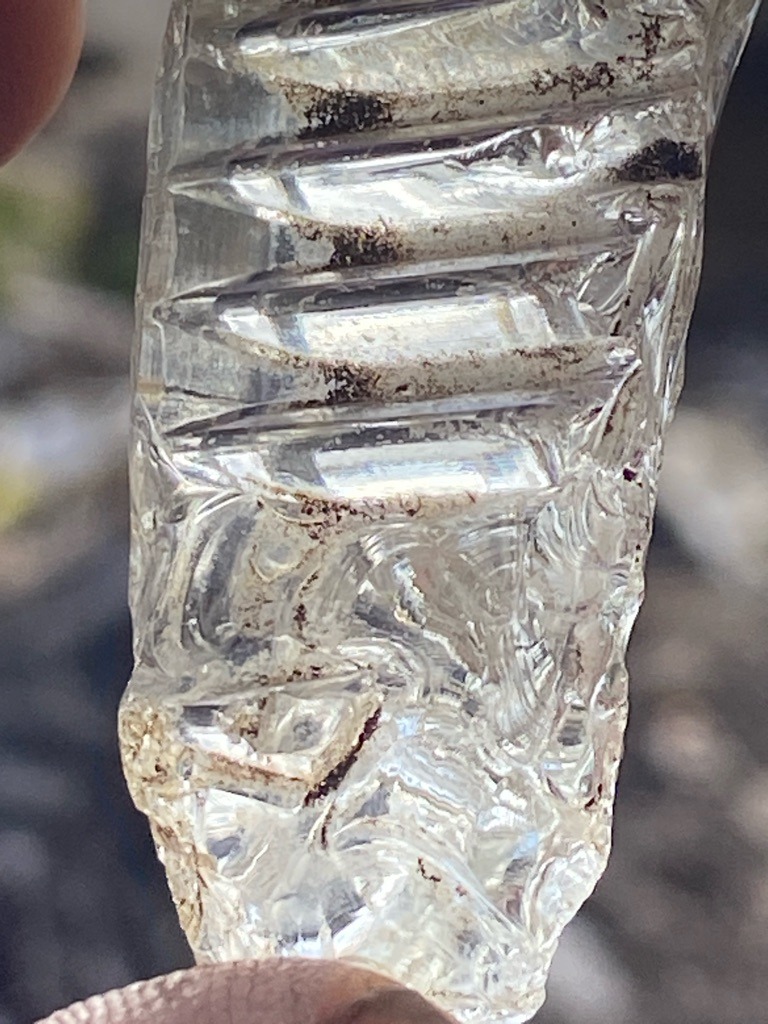 Not a diamond; this object appears to be a piece of heavily worked or carved natural quartz crystal or glass with conchoidal fractures and industrial/decorative facets, showing trapped dirt and debris in the grooves. - Irregular carved or fractured fragment