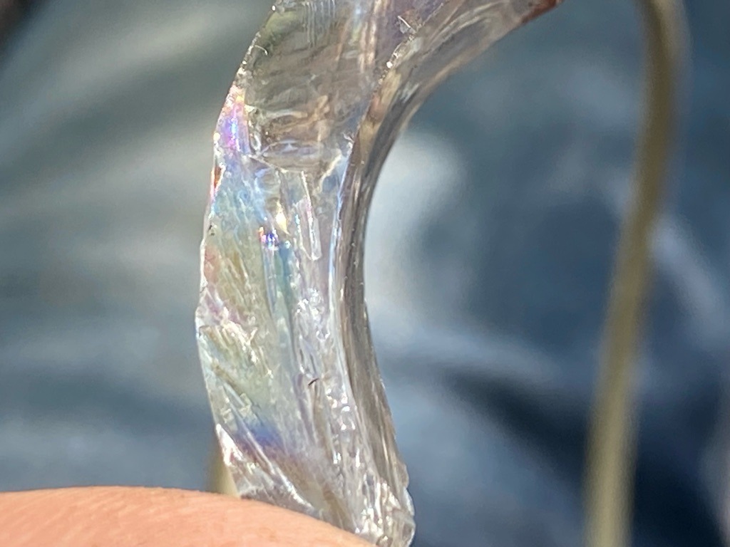 The image shows a substance that is not a diamond. It appears to be a curved piece of iridescent plastic, resin, or synthetic material with rough, uneven edges and internal crazing. - Irregular/Curved (appears to be a broken or molded curved fragment, not a traditional diamond cut)