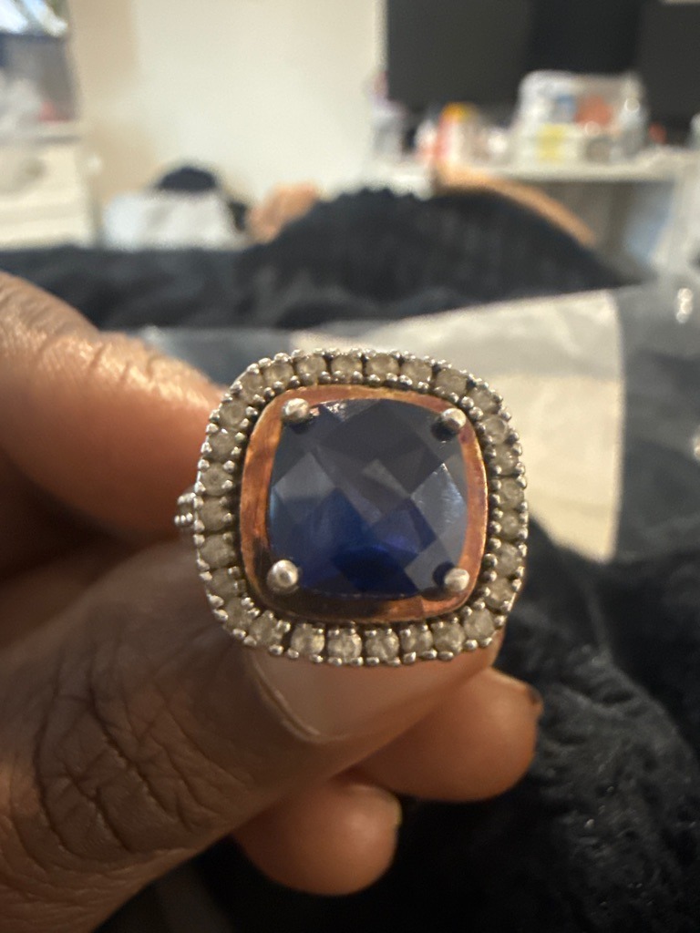 The central stone appears to be a natural or lab-created blue sapphire, surrounded by accent diamonds. - Cushion Modified Brilliant for the center blue stone, with round melee accent diamonds in the halo.