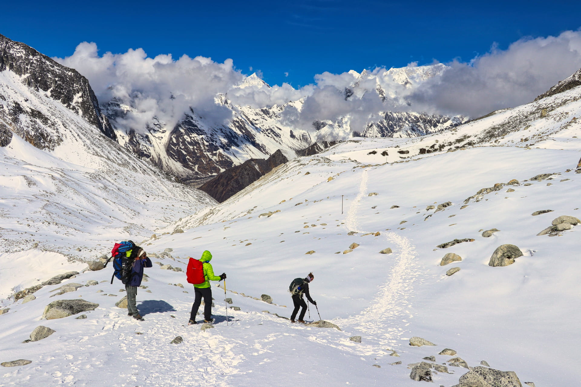 Troje pohodnikov s pisanimi nahrbtniki hodi po zasnežni hribovski poti na treku Manaslu