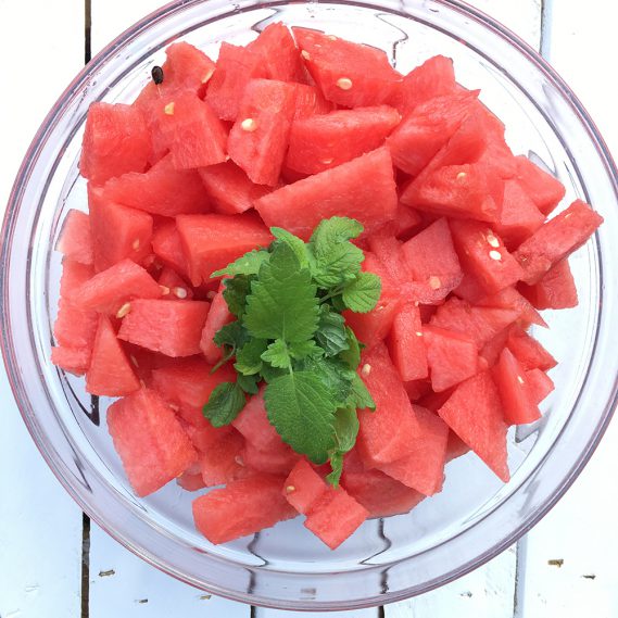 Melonenwürfel in Glasschale mit Zitronenmelisse auf hellem Holz Draufsicht