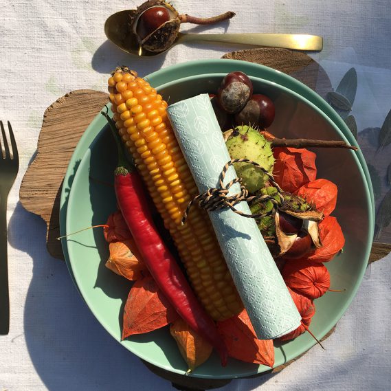 Herbstlich dekorierter Teller, mit Lampionblumen, Mais, Chillischote, goldenem Besteck und Kastanien