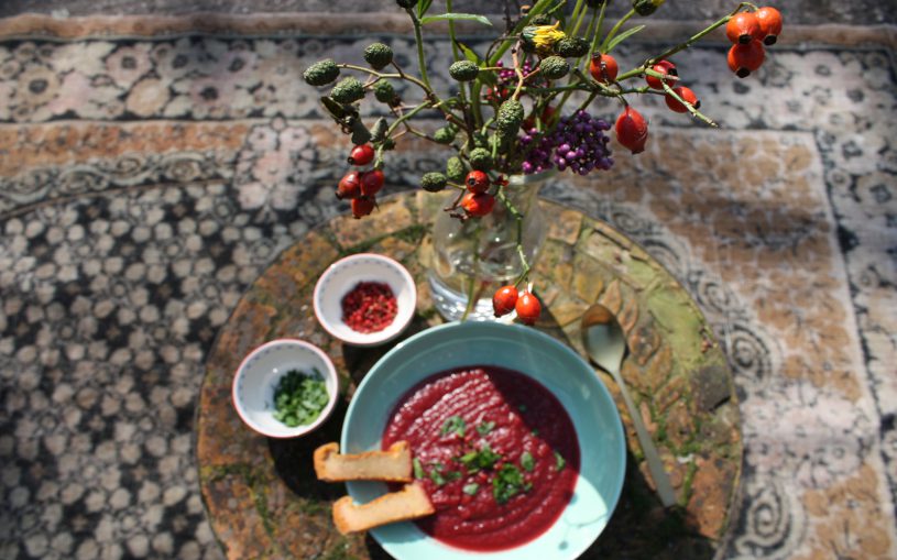 Rote Beete-Suppe auf grünem Teller, auf altem Gartentisch mit Herbststrauß und rosa Pfeffer und Petersilie, auf altem verblichenem Teppich