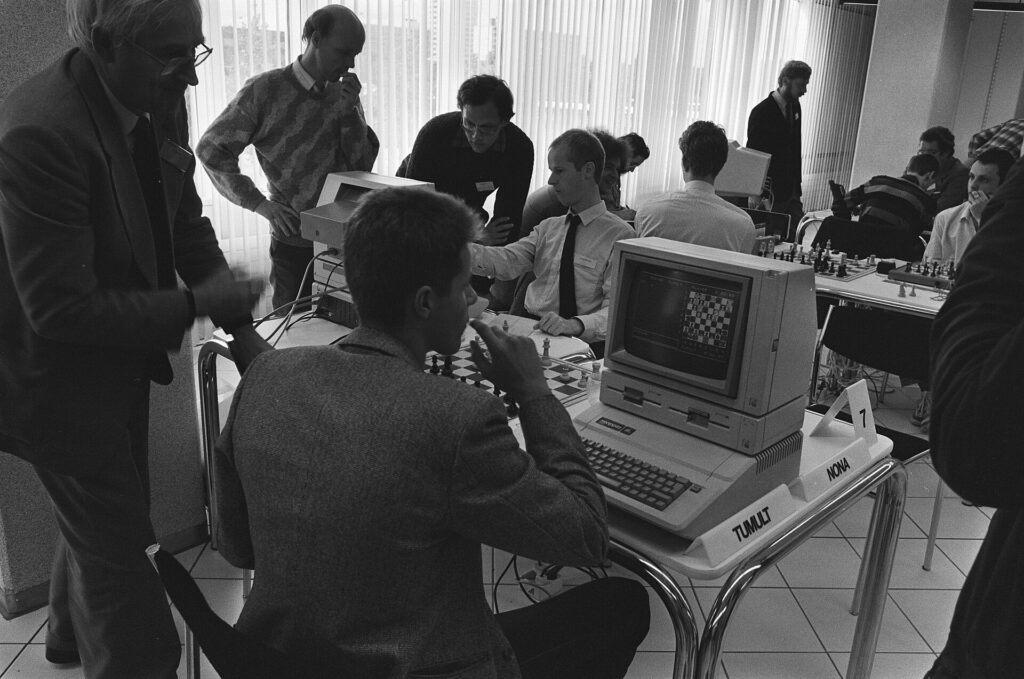 A photograph of an early computer chess competition with a room full of people, computers, and chess sets.