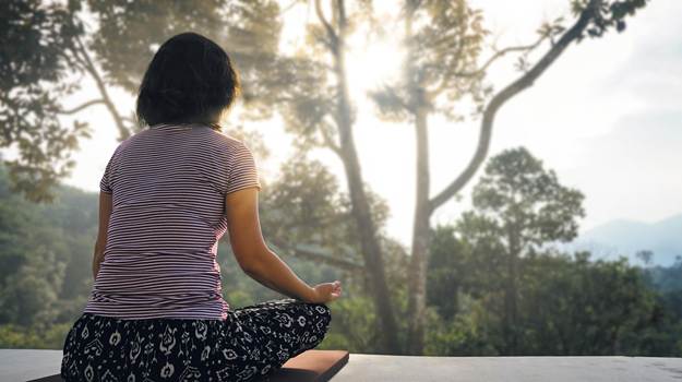A woman is seated in a mediation pose with her back to the camera, viewing a natural landscape