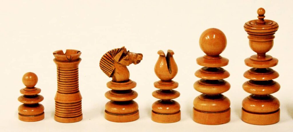 Six chess pieces from a St. George set are shown in a line, each piece has many carved details in a light colored wood