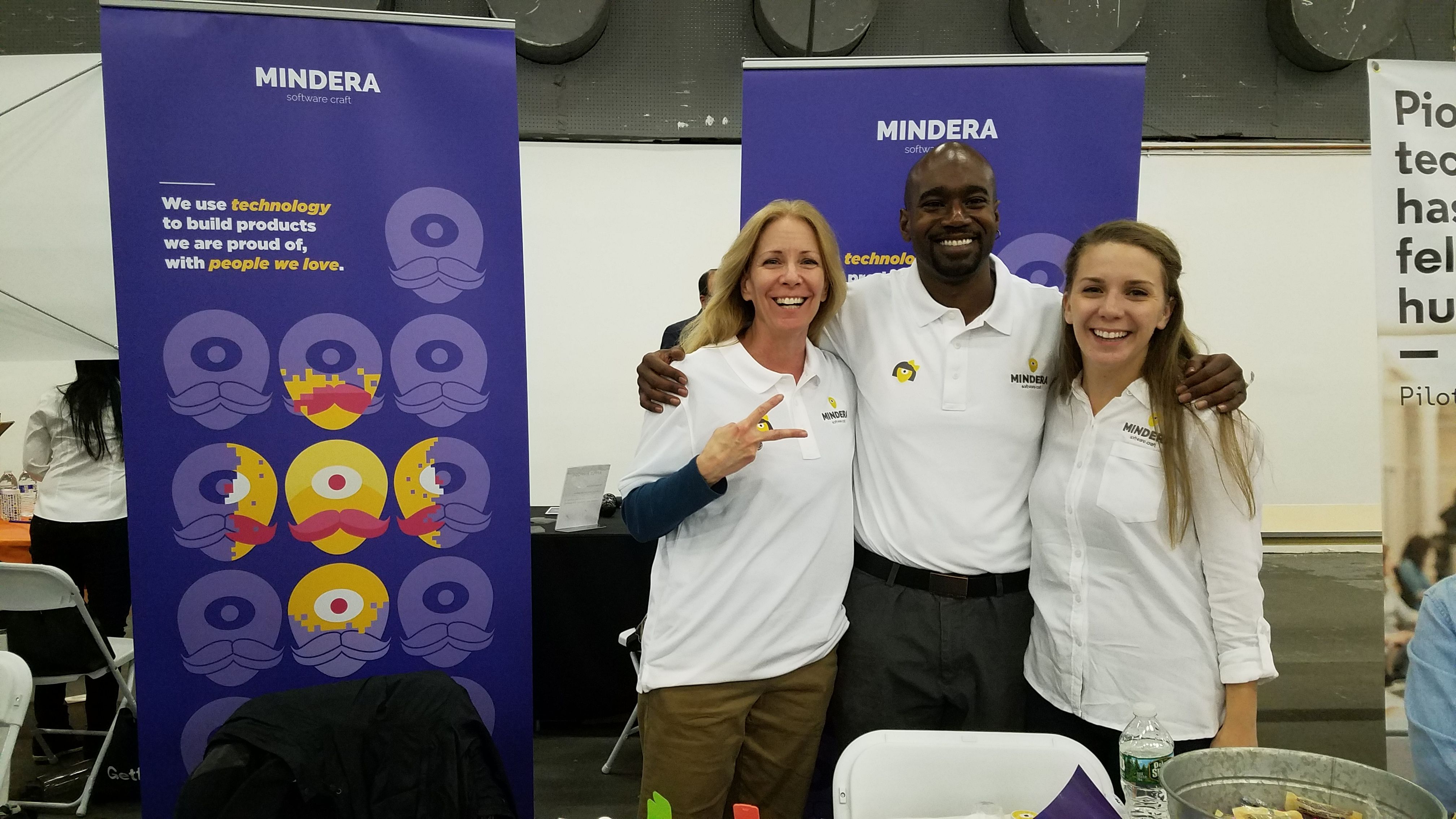 A male Minder stands in the middle of two female Minders as they pose in front of a purple sign that says "Mindera" in white text.