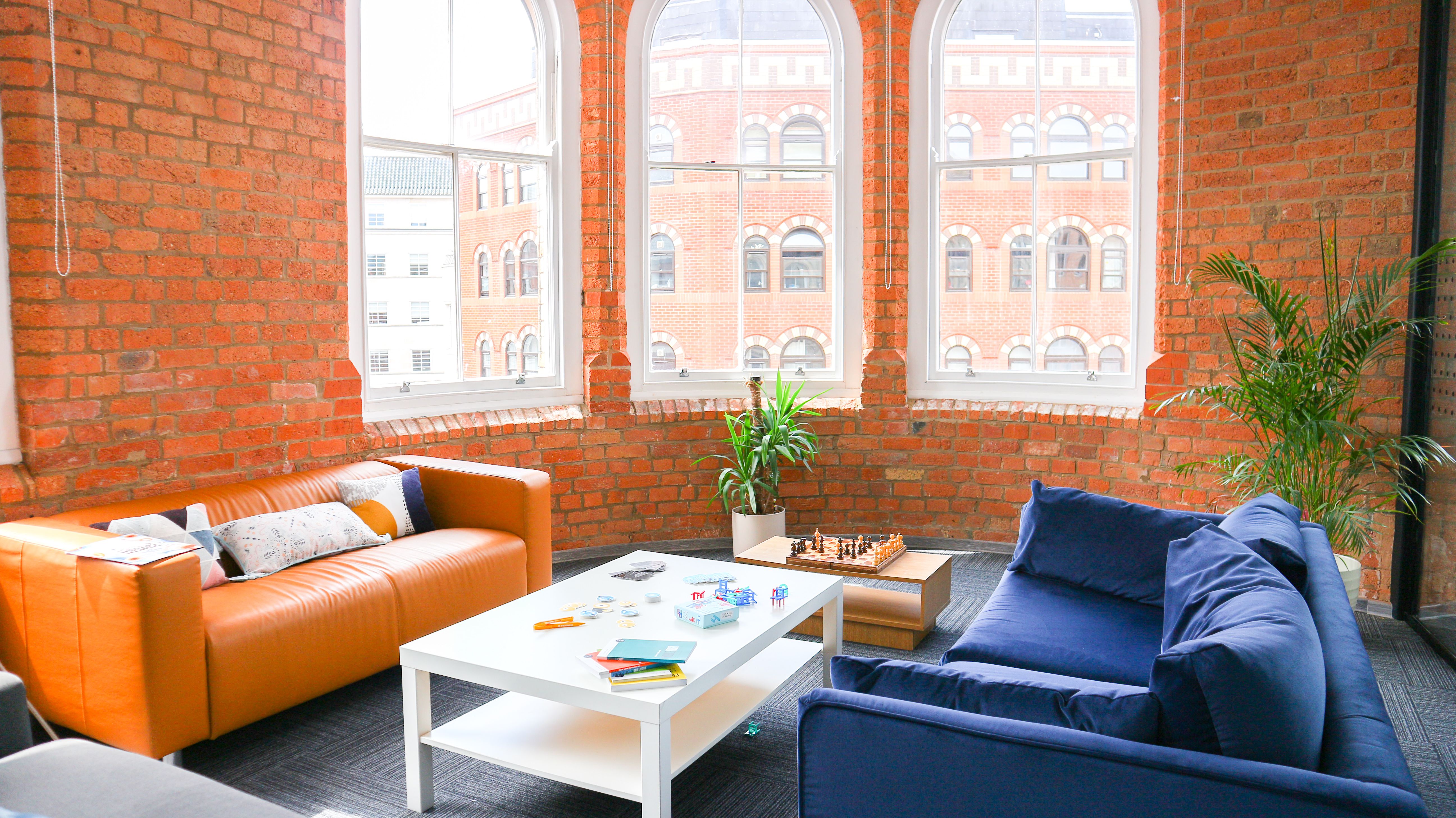 An empty orange sofa and an empty blue sofa, with a white table between them and a wall of orange bricks behind them, in the Mindera - UK office. 