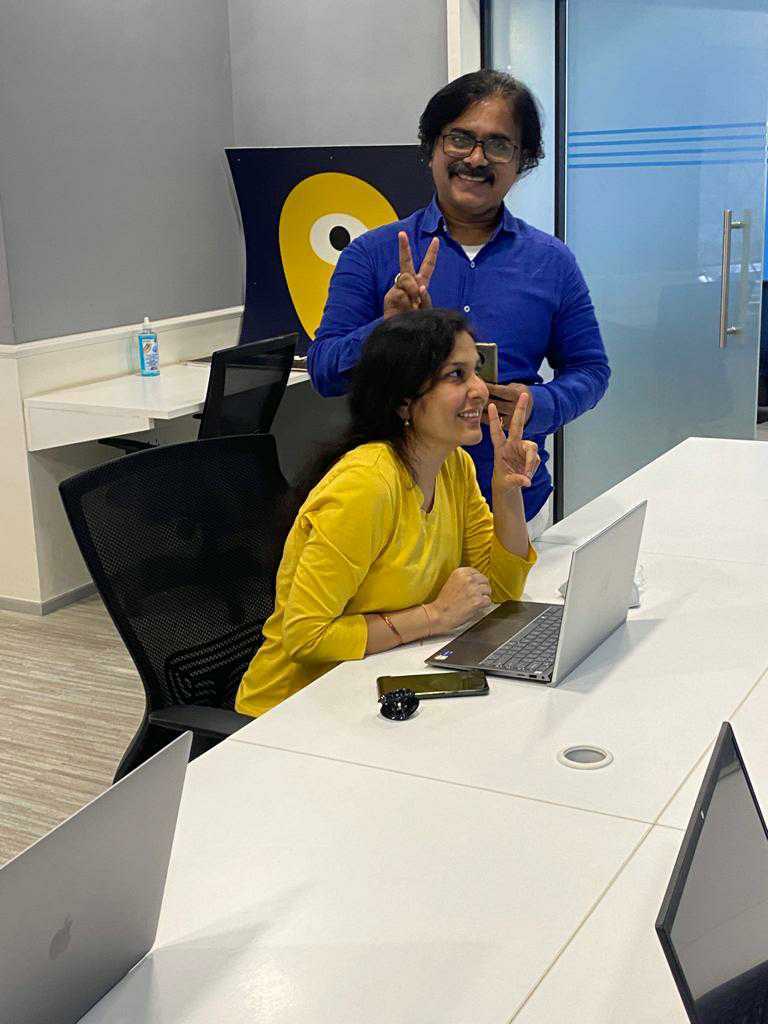 A female Minder in a yellow blouse and a male Minder in a blue shirt both give the peace sign with their fingers as they stand in front of the Mindera logo.