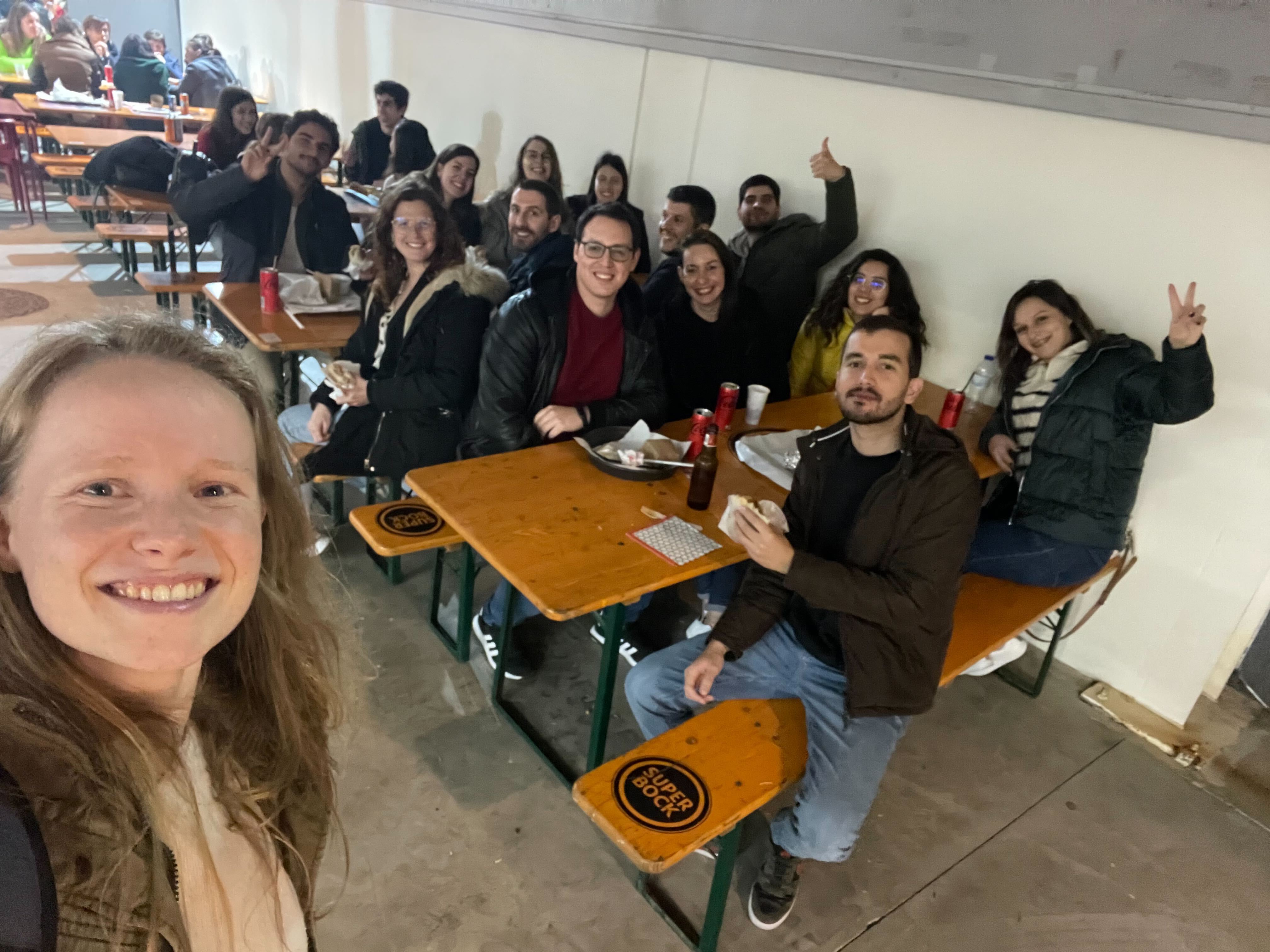 A Minder takes a selfie that shows a group of Minders sat at tables in the background, having food and drinks as they discuss the Mindera About Us page. 