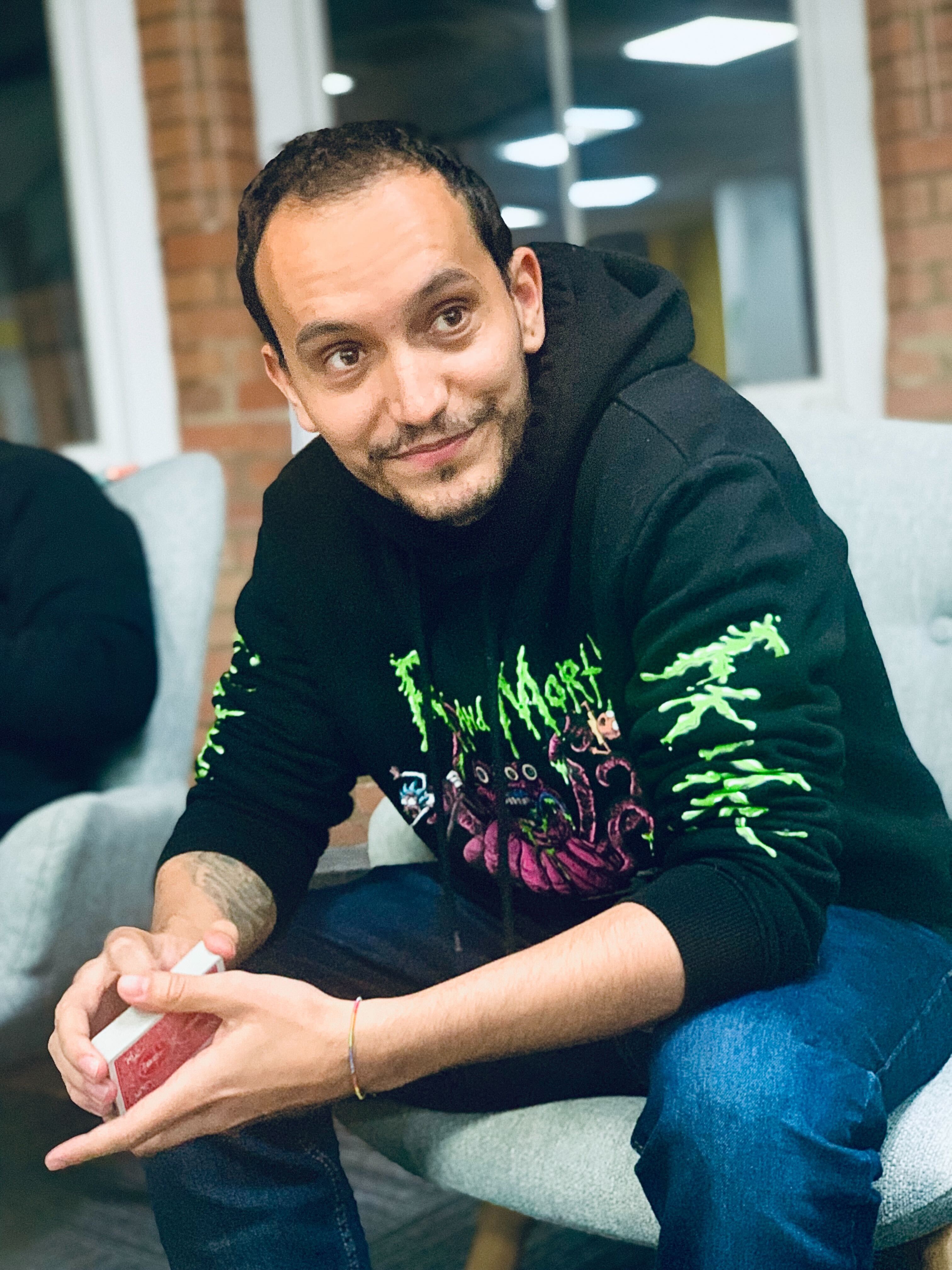 A photo of Daniel Andrade, a Recruitment Strategist for software engineering company Mindera, wearing a black hoodie and blue jeans while he holds a back of playing cards.