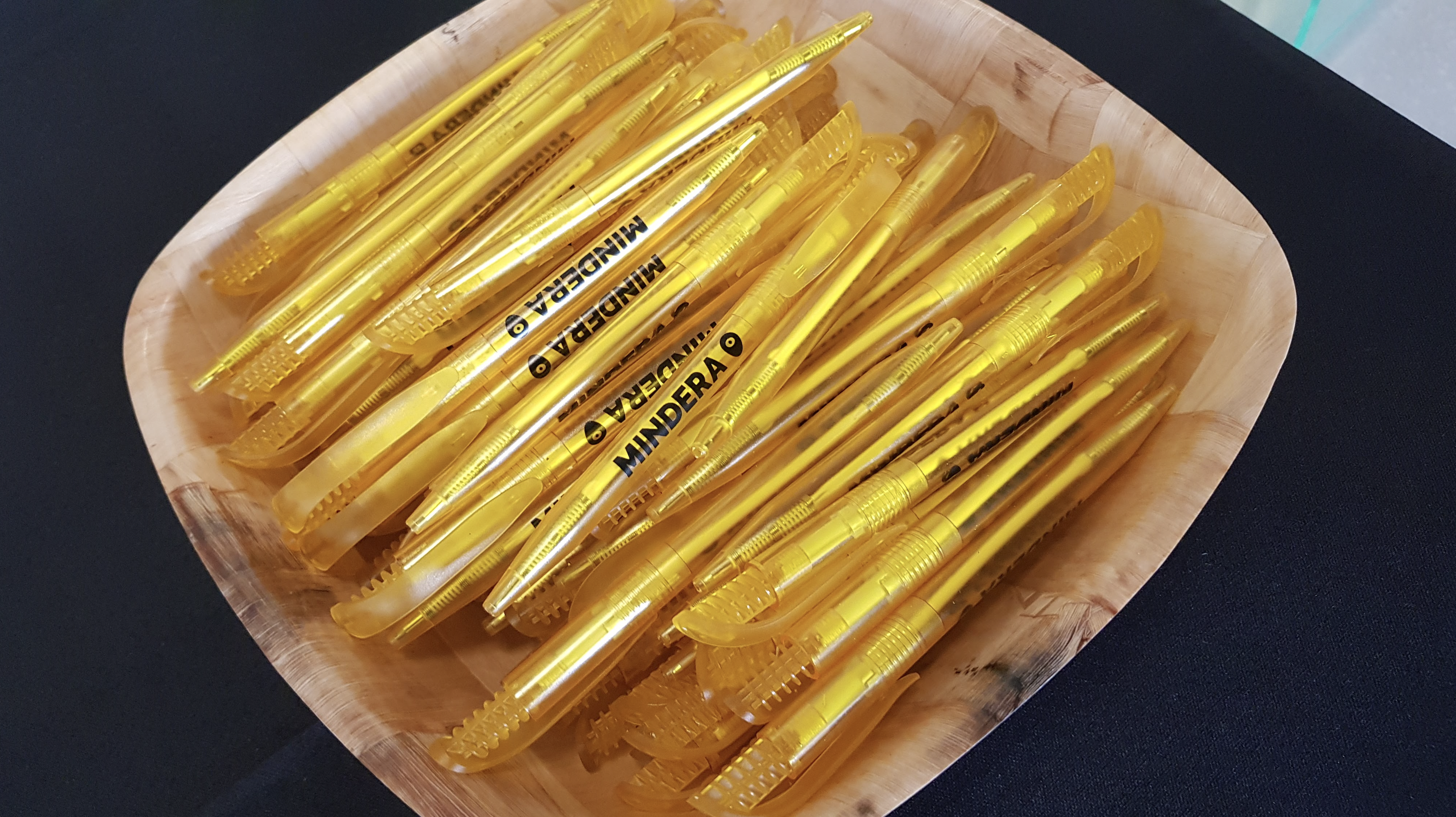 A bunch of yellow pens with the black logo and black name of software engineering company Mindera on them rest in a wooden bowl. 