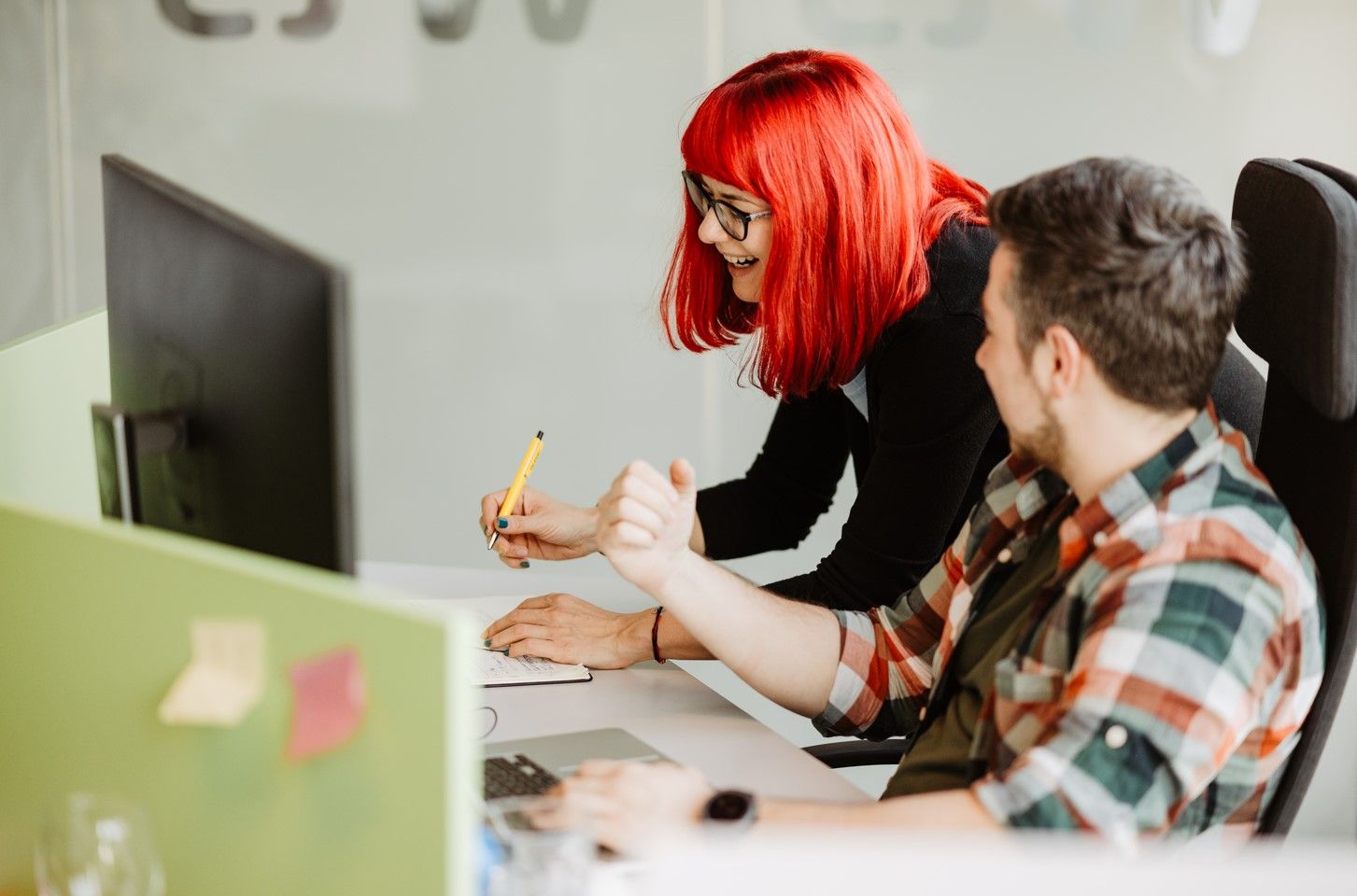 A female Minder with red hair and a male Minder with brown hair sit in profile in front of a monitor and laptop..