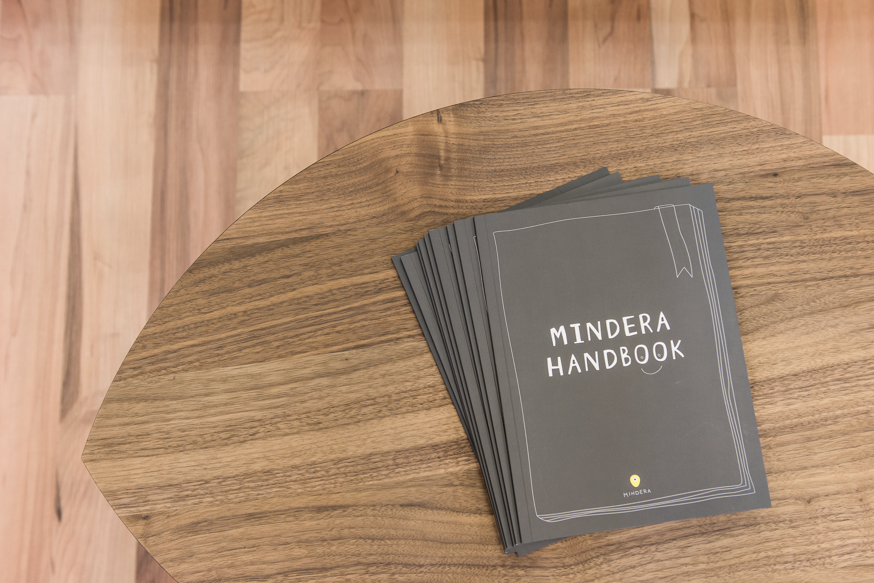 Grey copies of the handbook of software engineering company Mindera are piled on top of each other on a wooden table, with Mindera Handbook written on the handbooks in white text. 