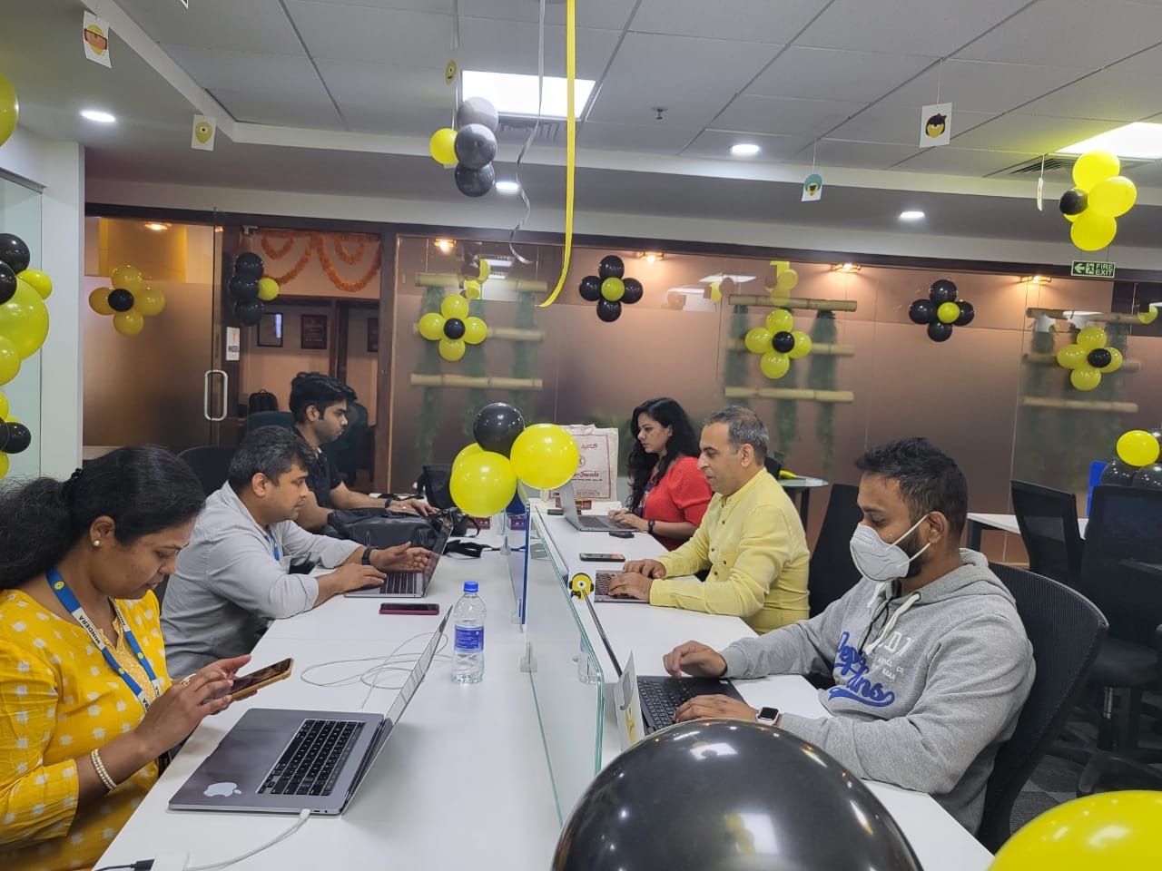 Six Minders are working in their Macbooks in an office decorated with yellow and black balloons.