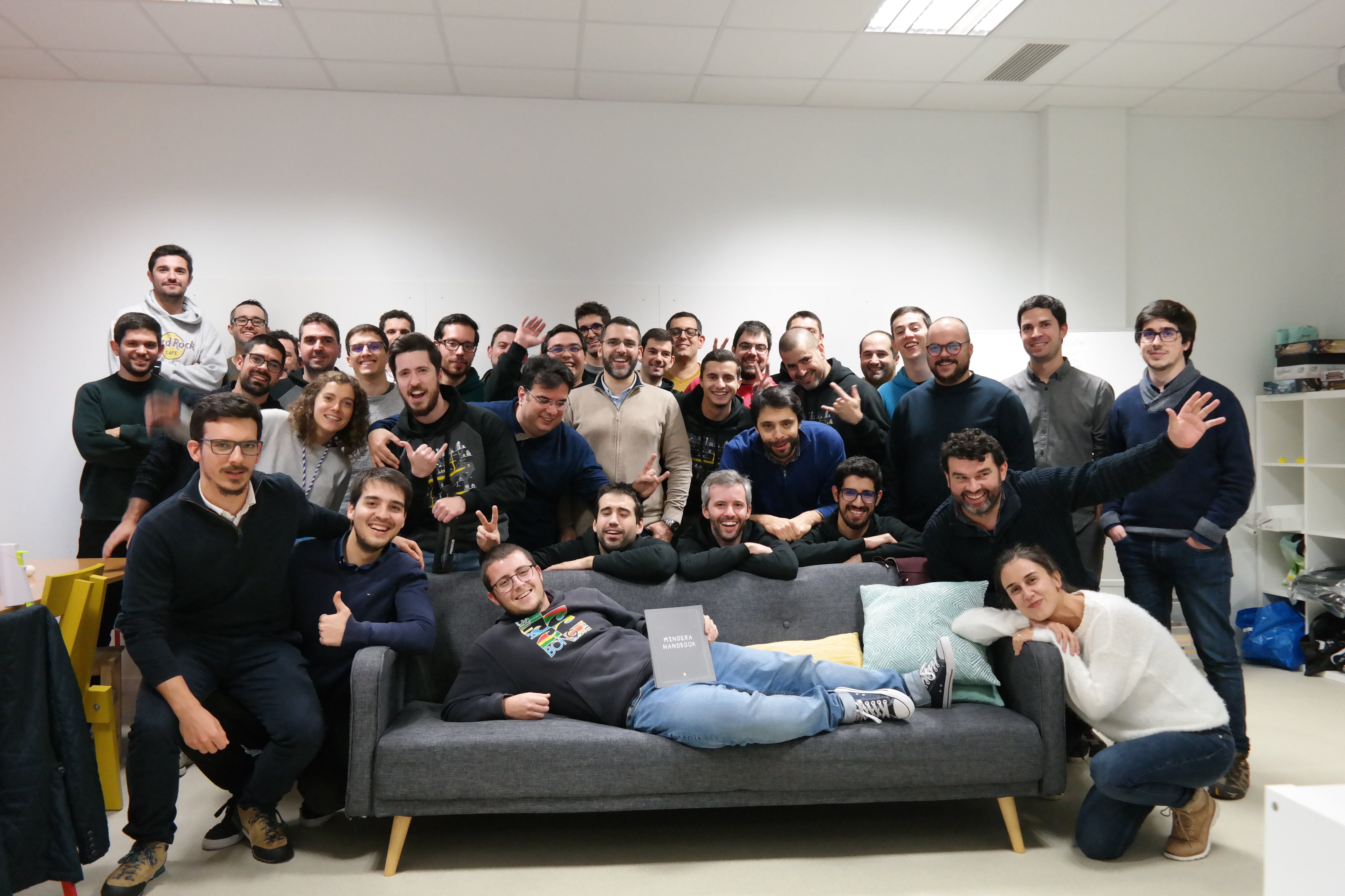 A group of many Minders pose for the camera, with some lying and leaning on a sofa at the centre of the picture, in the office of software engineering company Mindera.  