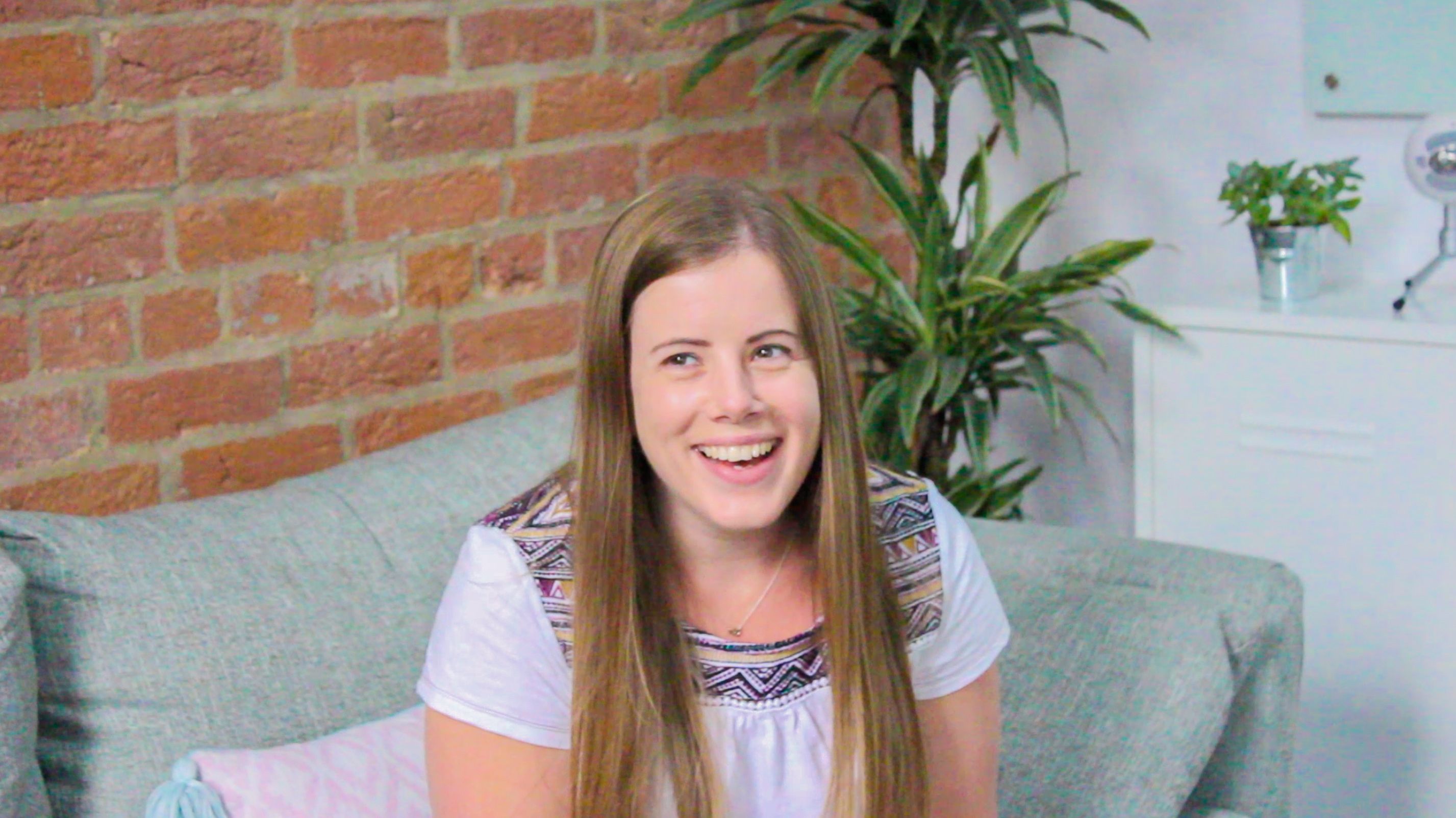 A photo of Gemma Forrest, a People Operations professional who works for software engineering company Mindera, smiling while she sits on a grey/green sofa with a plant in the background. 