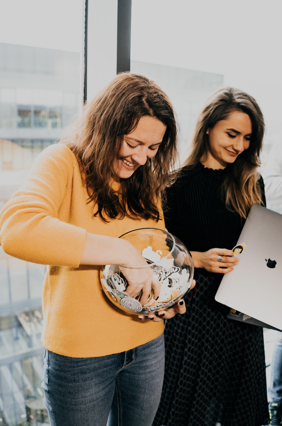 Two female Minders are stood in front of a window, one holds a glass bowl and the other holds a grey MacBook.