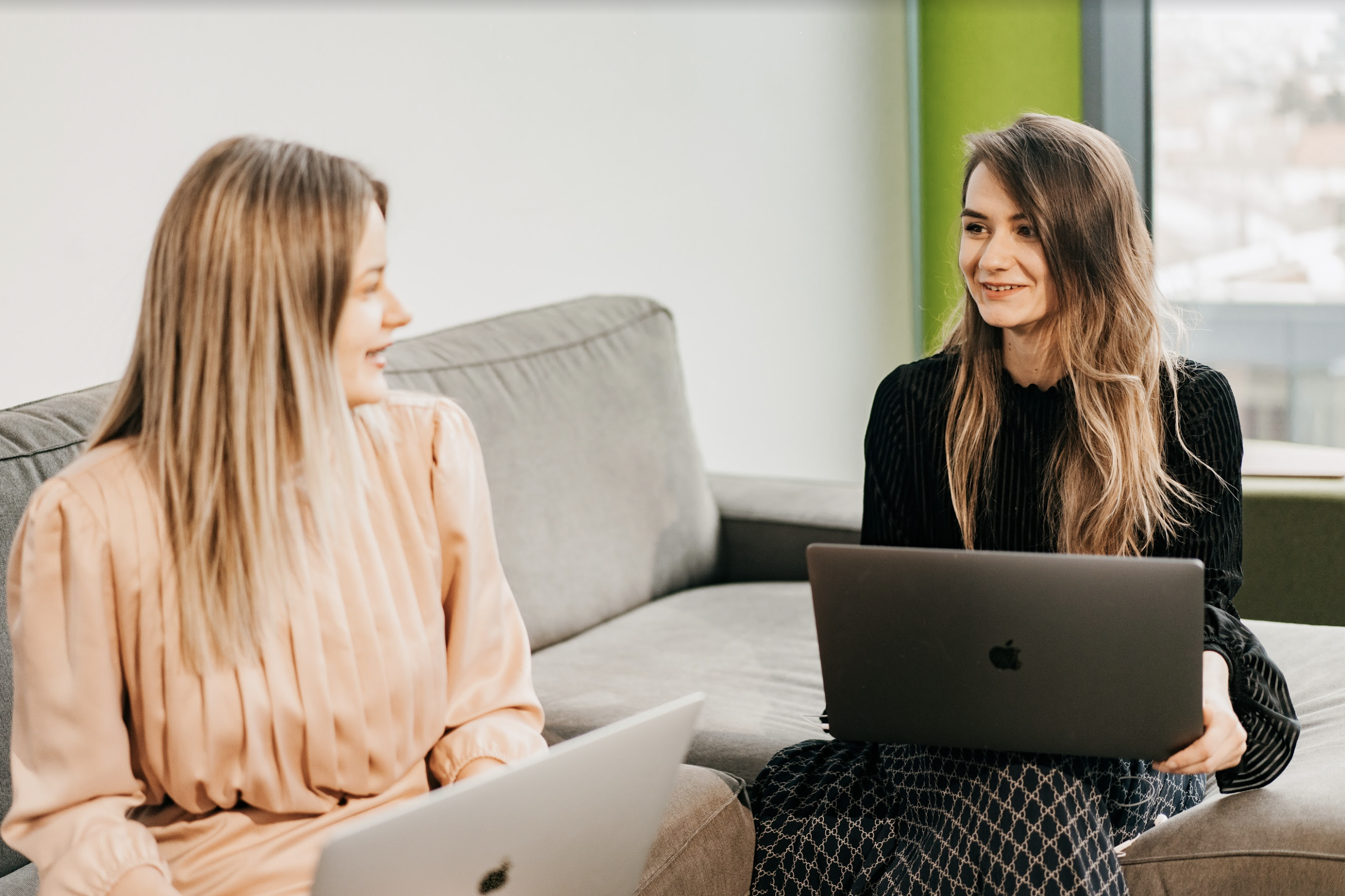 Two female Minders, each holding a MacBook, are sat on a light grey couch in the Minder Romania office. 