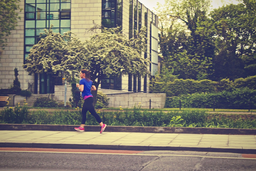 A woman runs in profile on the pavement, with trees and buildings to her right. 