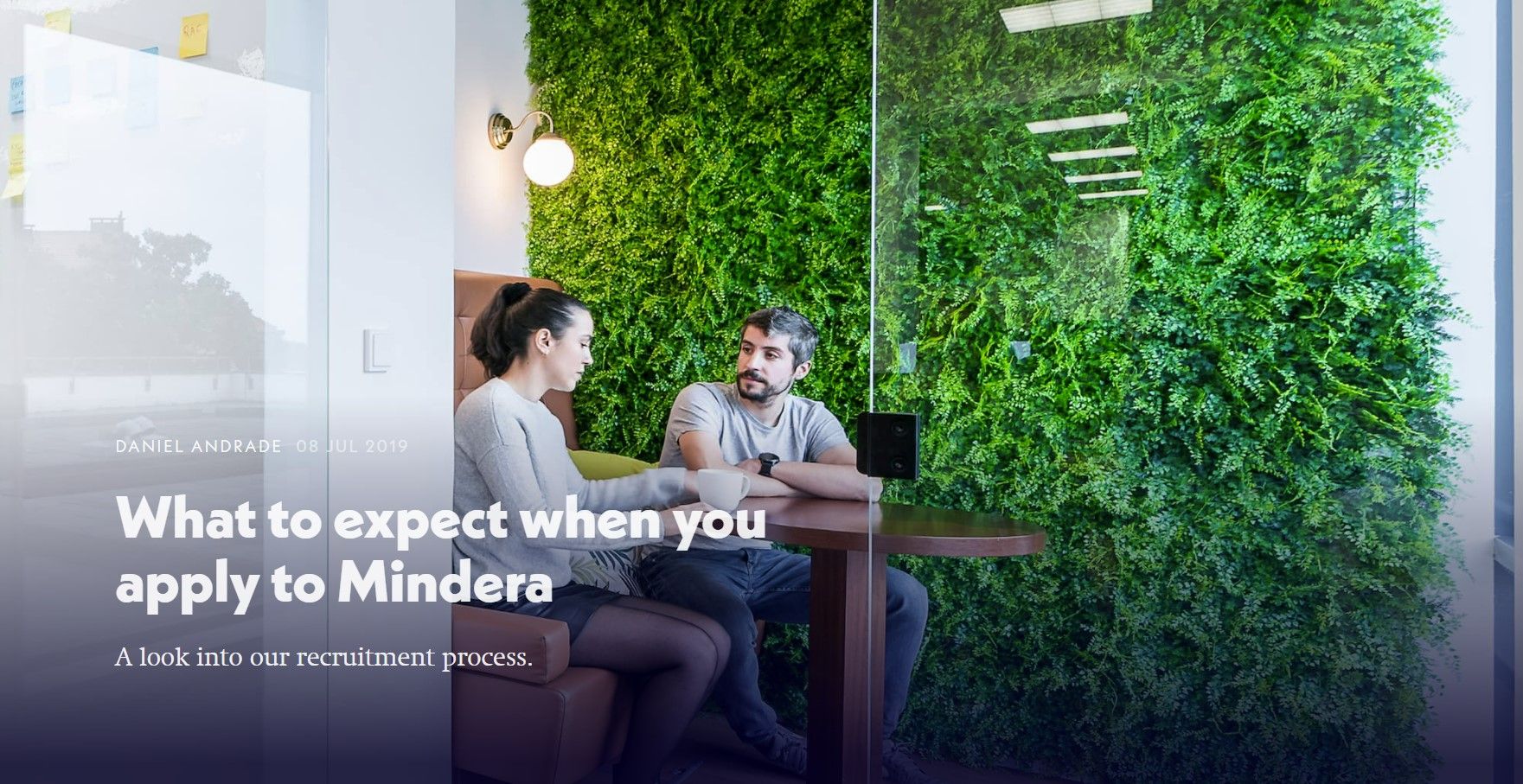 A female Minder and a male Minder sit at a table, with a window to their left and Astroturf wall to their right, as they discuss what to expect when you join Mindera.