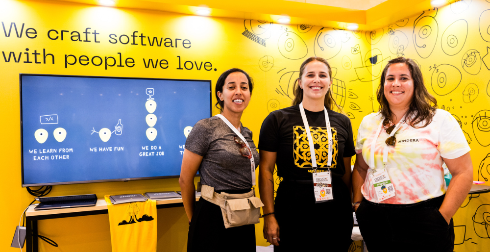 3 Minders standing in our Booth at Devoxx Morocco. The background is the Mindera yellow with our Minder logo patterned on. A TV screen sits below our slogan on the wall.
