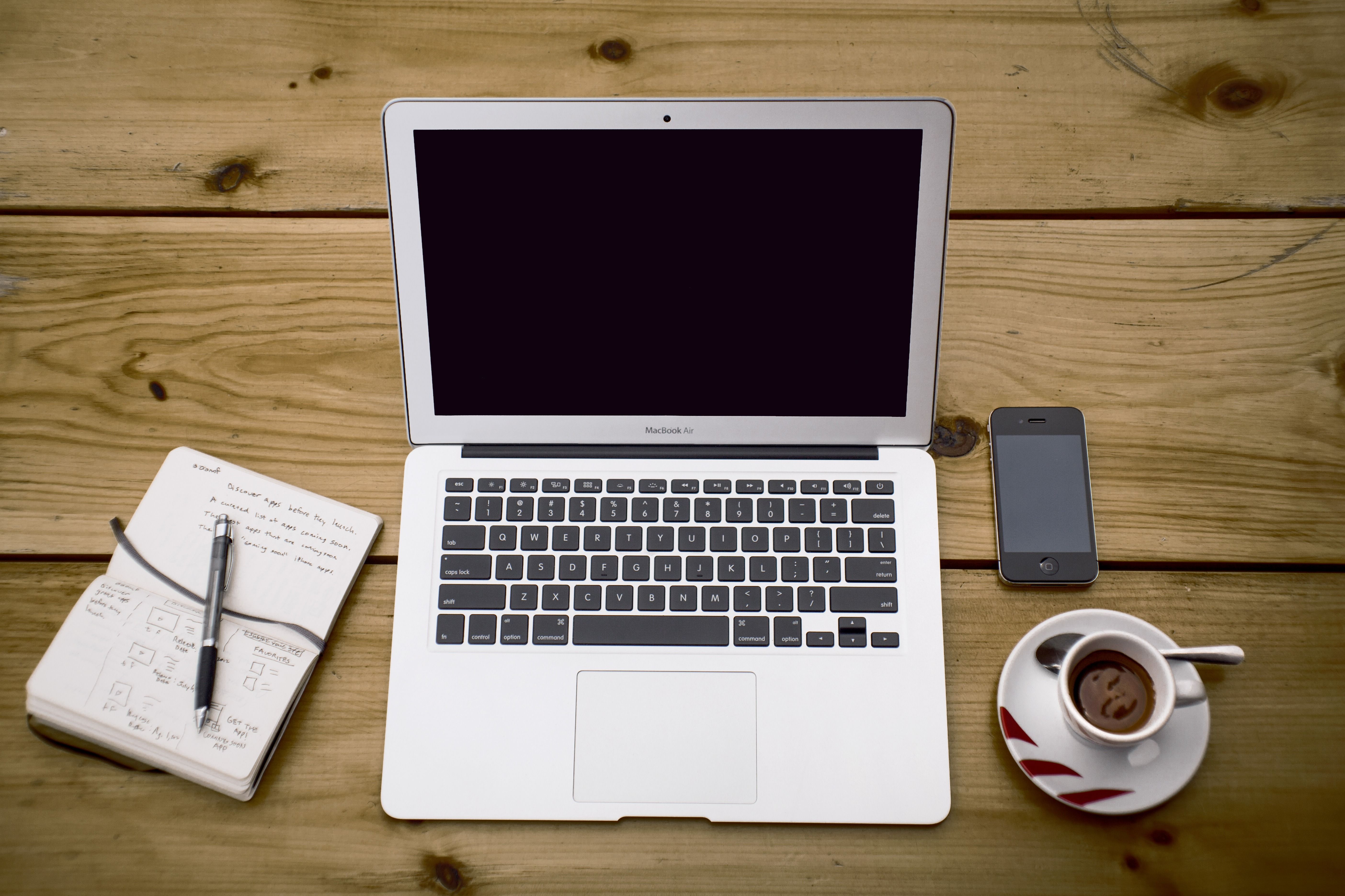 A MacBook rests on a wooden table, with a writing notepad and pen to its left and a iPhone and cup of coffee to its right.