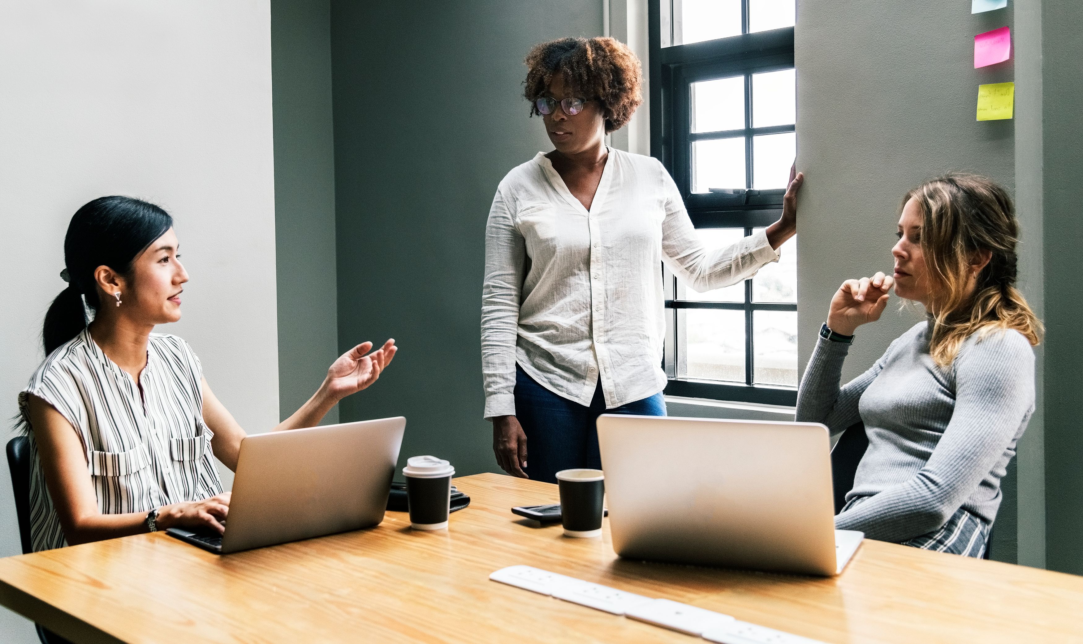 Two women sit at a table in front if their laptops as a woman gives a talk about women in tech.