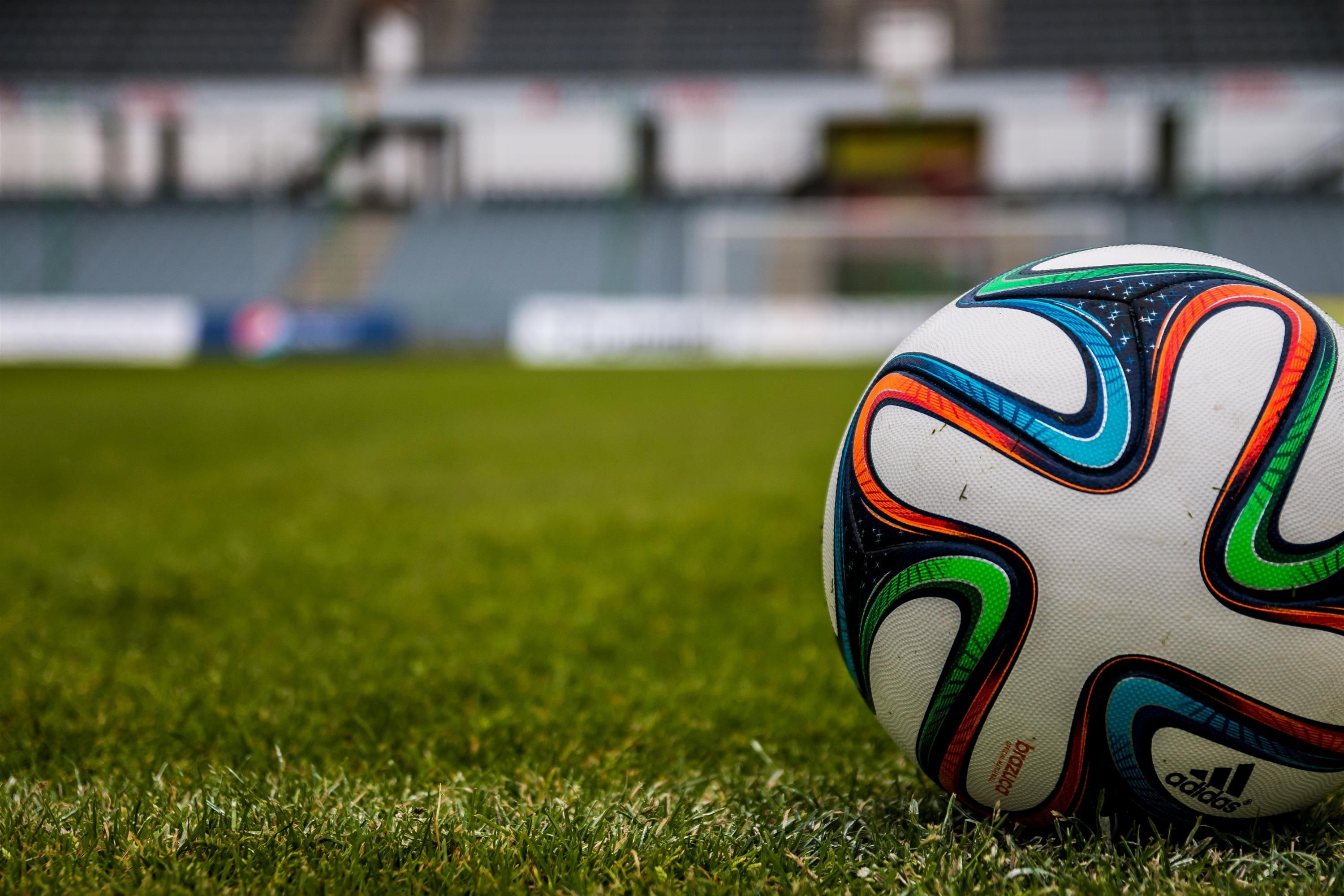 An Adidas football rests on grass, with a Mindera - United Kingdom Minder just out of view but ready to kick the ball. 