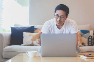 Asian attractive man with eyeglasses sitting couch home working laptop