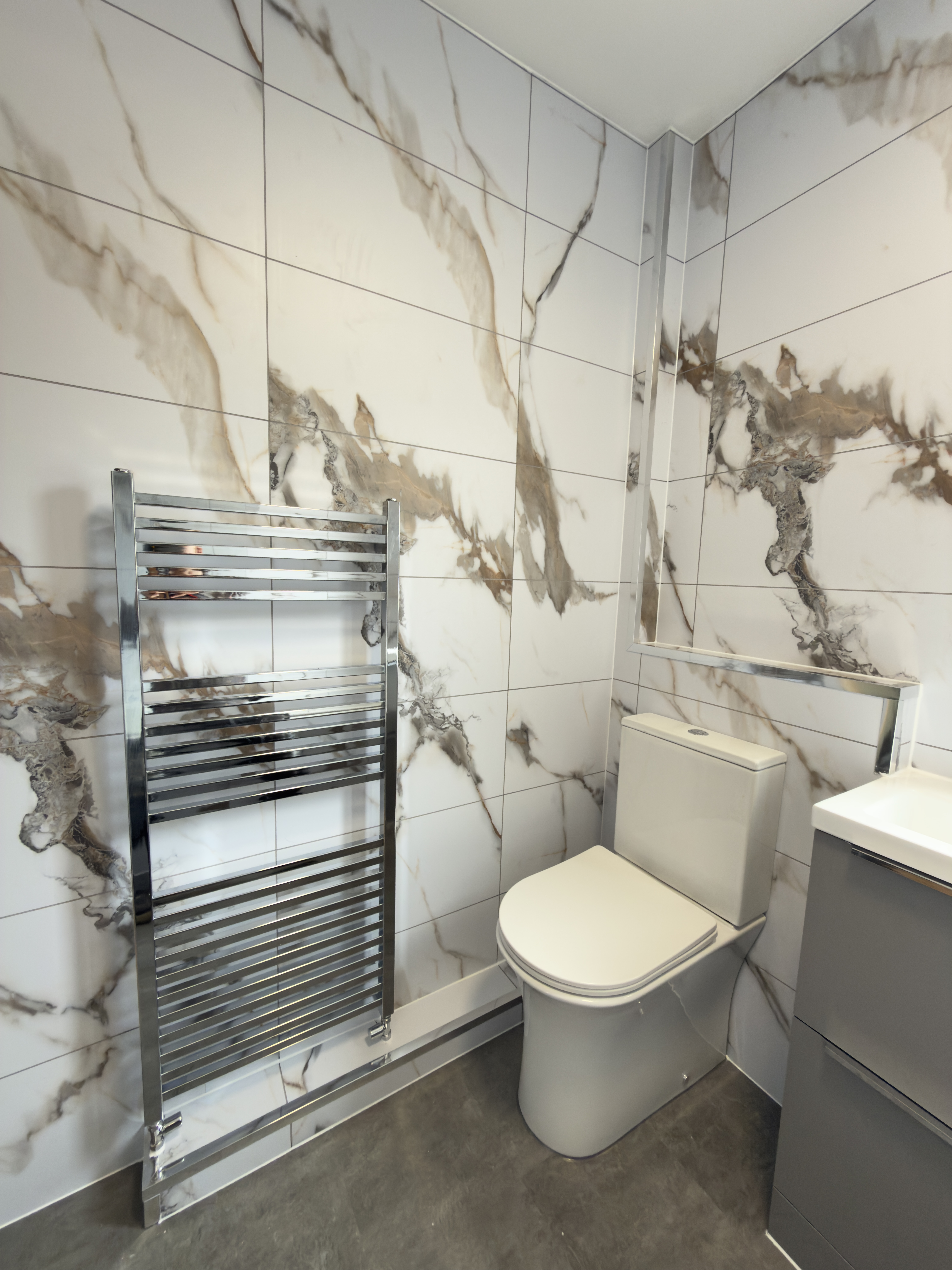 Chrome heated towel radiator and toilet against a backdrop of Calcutta Gold wall boards.