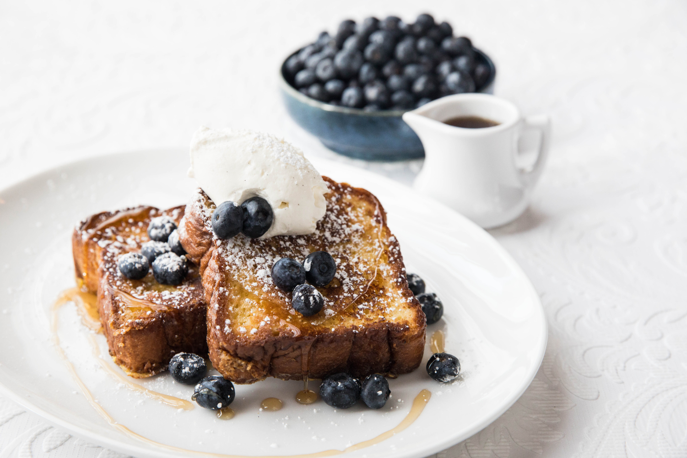 Mon Ami Gabi's French Toast with blueberries and chantilly cream with a bowl of fresh blueberries and a cup of maple syrup next to it.