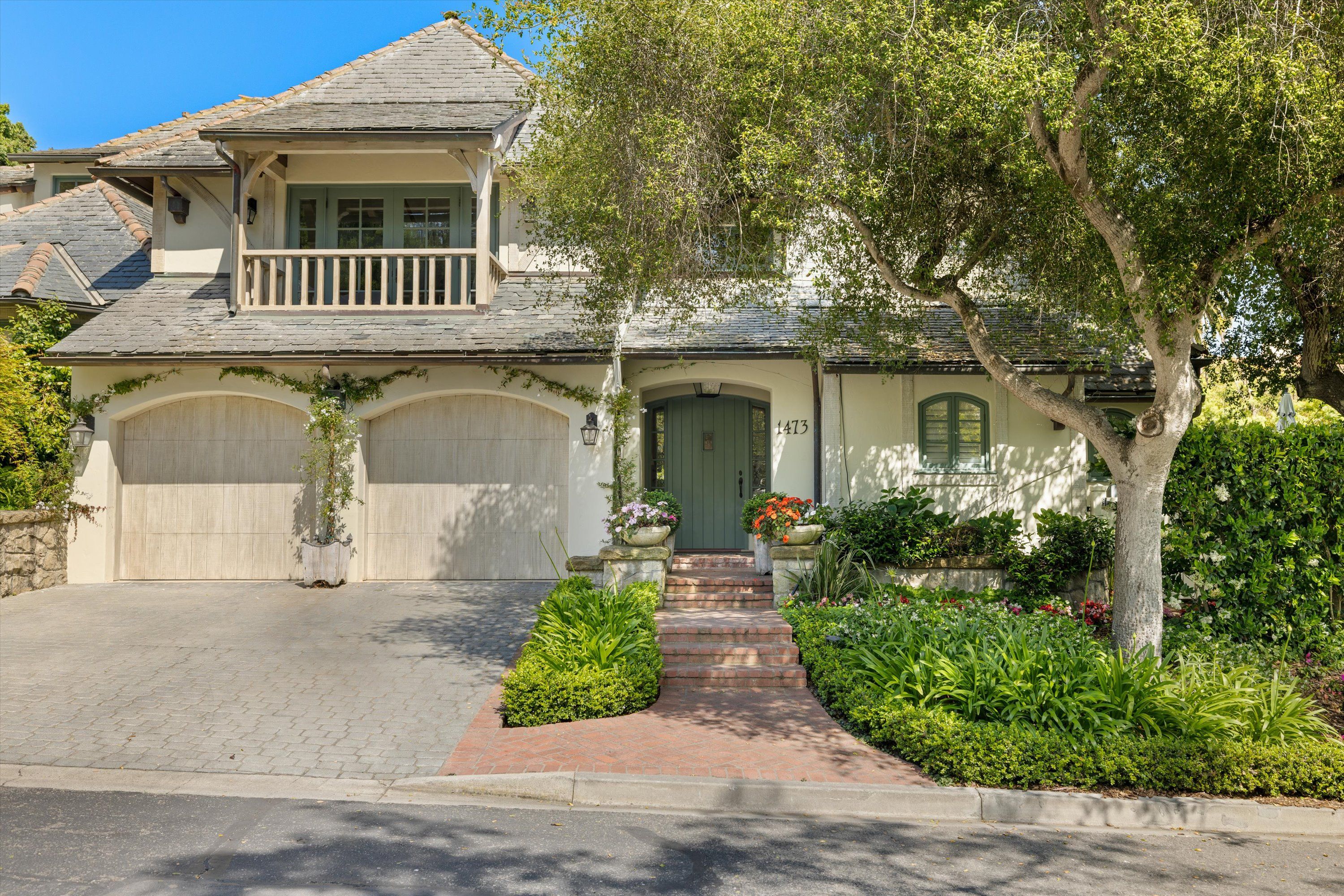 The front of a luxury French country style bungalow with a balcony above its 2-car garage, and a tree in the front.