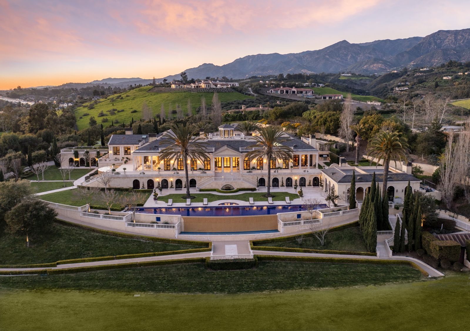 A magnificent estate with mountains and the sunset in the background, with the estates massive g rounds and lush landscaping in the foreground