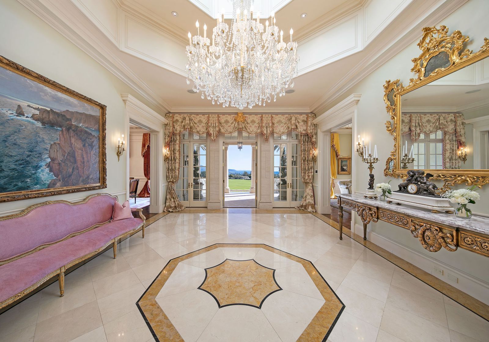 The elegant Palladian-style foyer of a mansion, with crystal chandelier overhead, a highly polished marble floor, a gilded sideboard and mirror, long pink Victorian-style sofa, and th ocean in the background through draperied French doors