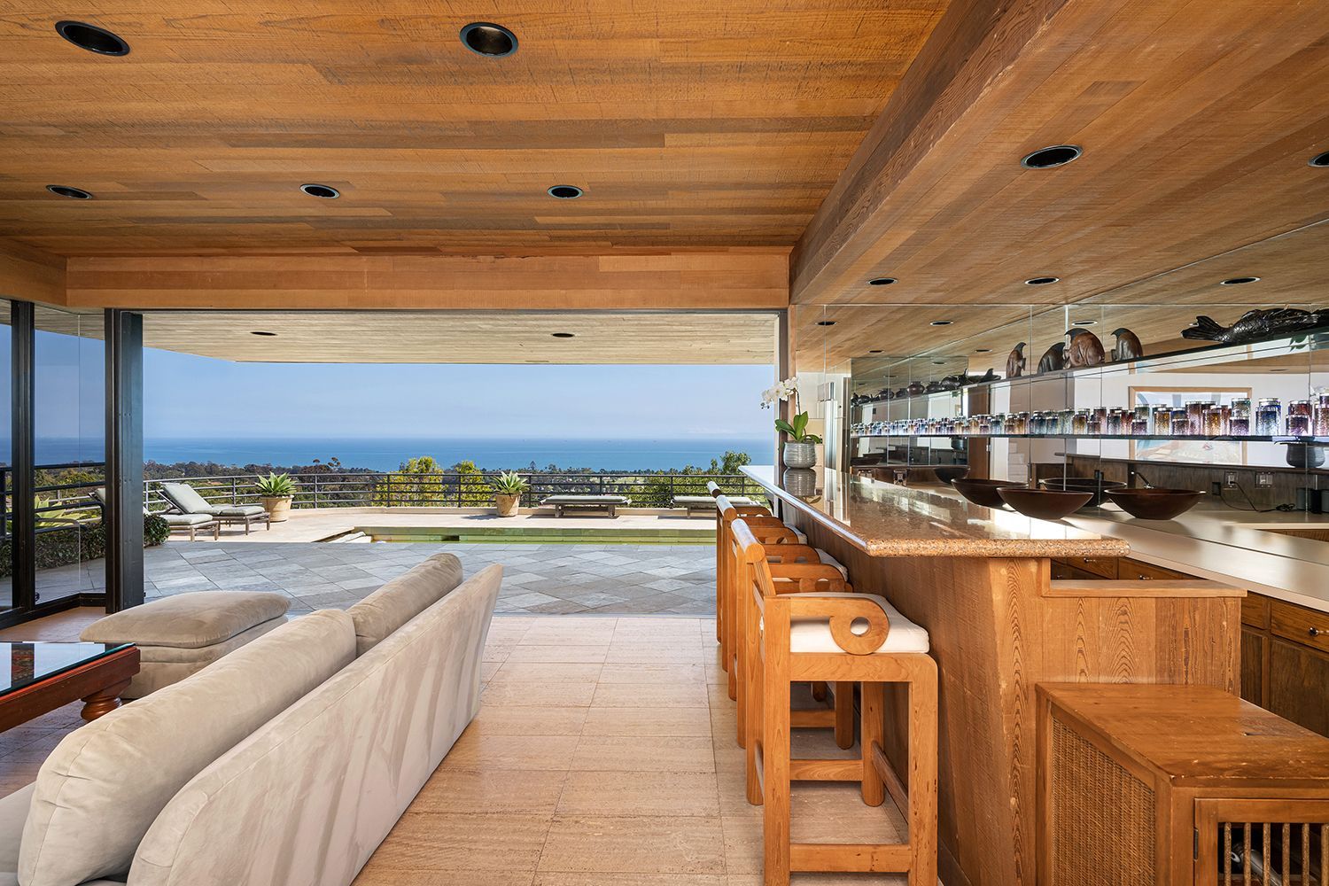 A room with wood plank ceiling, a bar and large sofa opening seamlessly to a pool, terrace, and ocean view