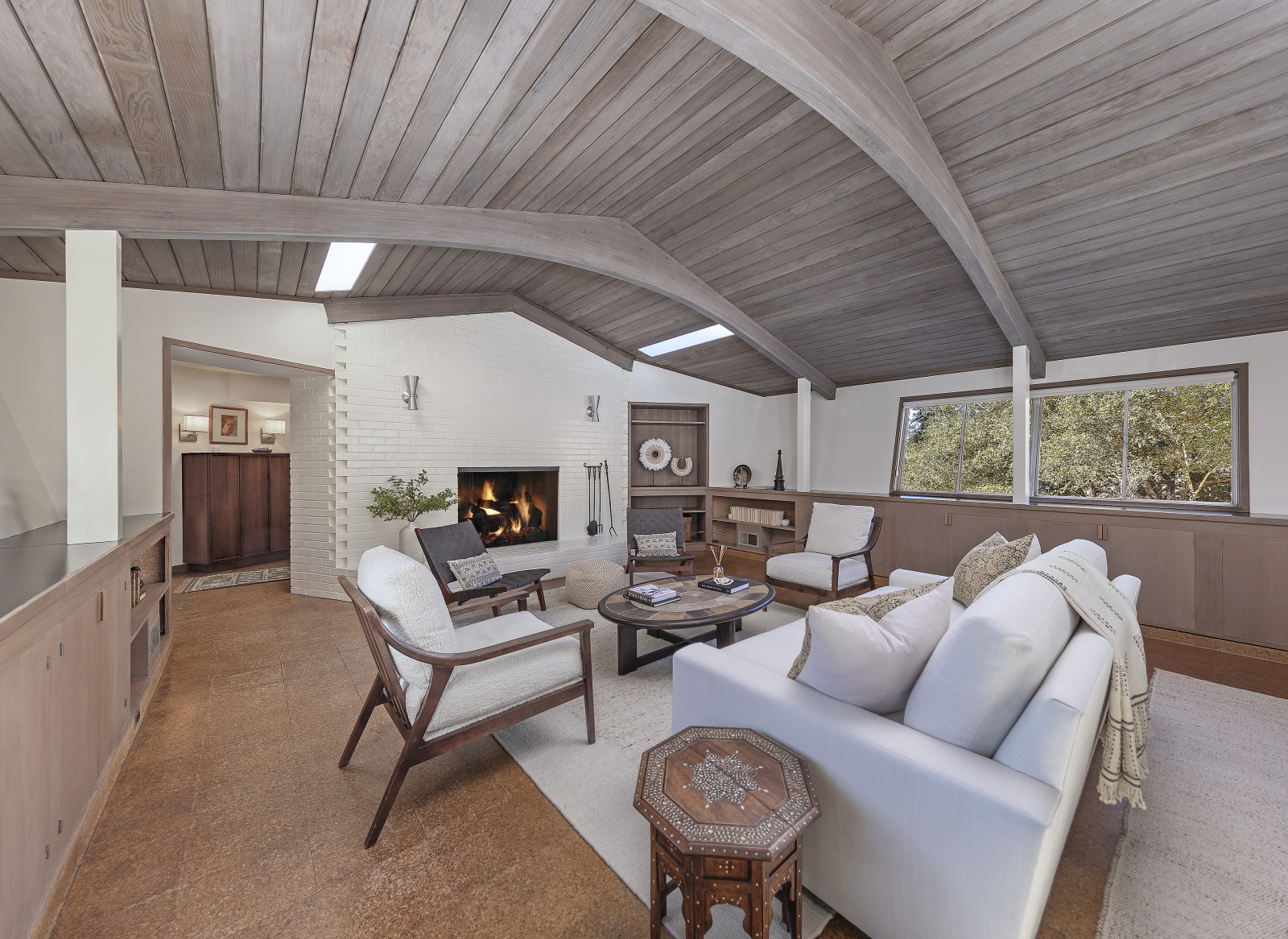 Montecito living room with vaulted wood beam ceiling, white brick fireplace, built-in cabinetry, neutral furnishings, and large windows with natural light.
