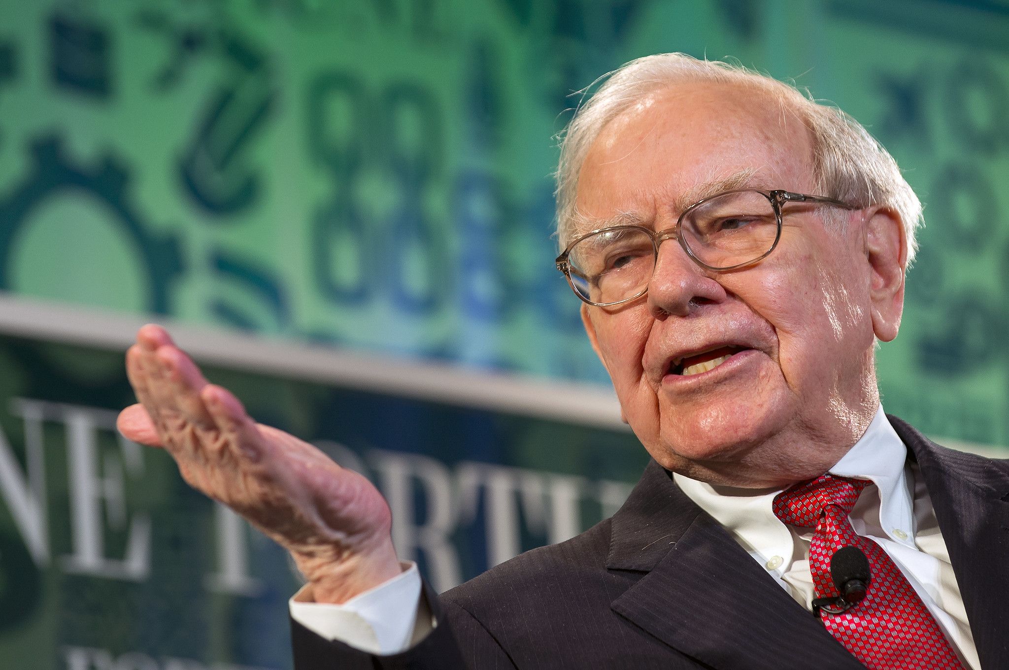 Warren Buffet in a suit and tie during a speech in front of a green screen.