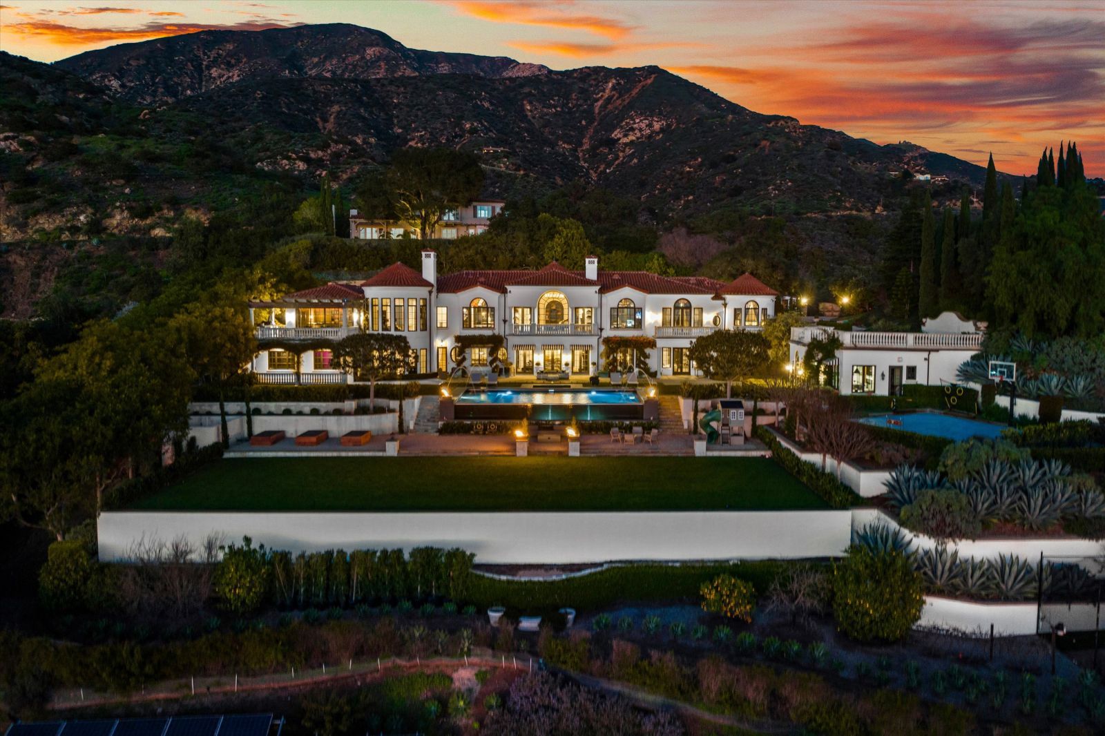 The front view of a multi-level Montecito Estate at sunset, lit up from withinwith a red tiled roof and white stucco exterior surrounded by lush vegetation and the mountains in the background