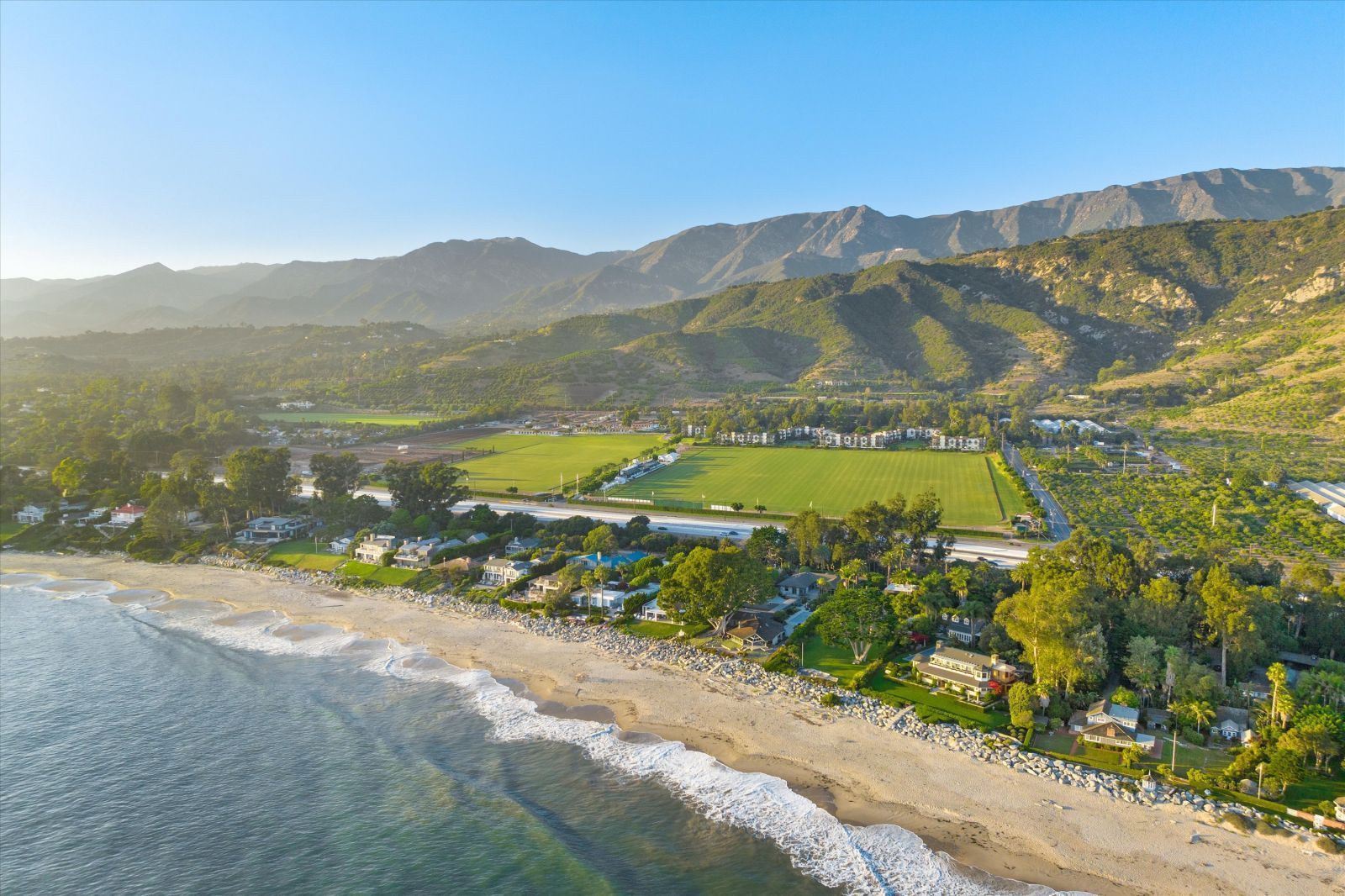 A drone shot of the Carpinteria coastline, with blue ocean, white surf, and sandy beach, along with homes, the Santa barbara Polo Club, and mountains in the background.