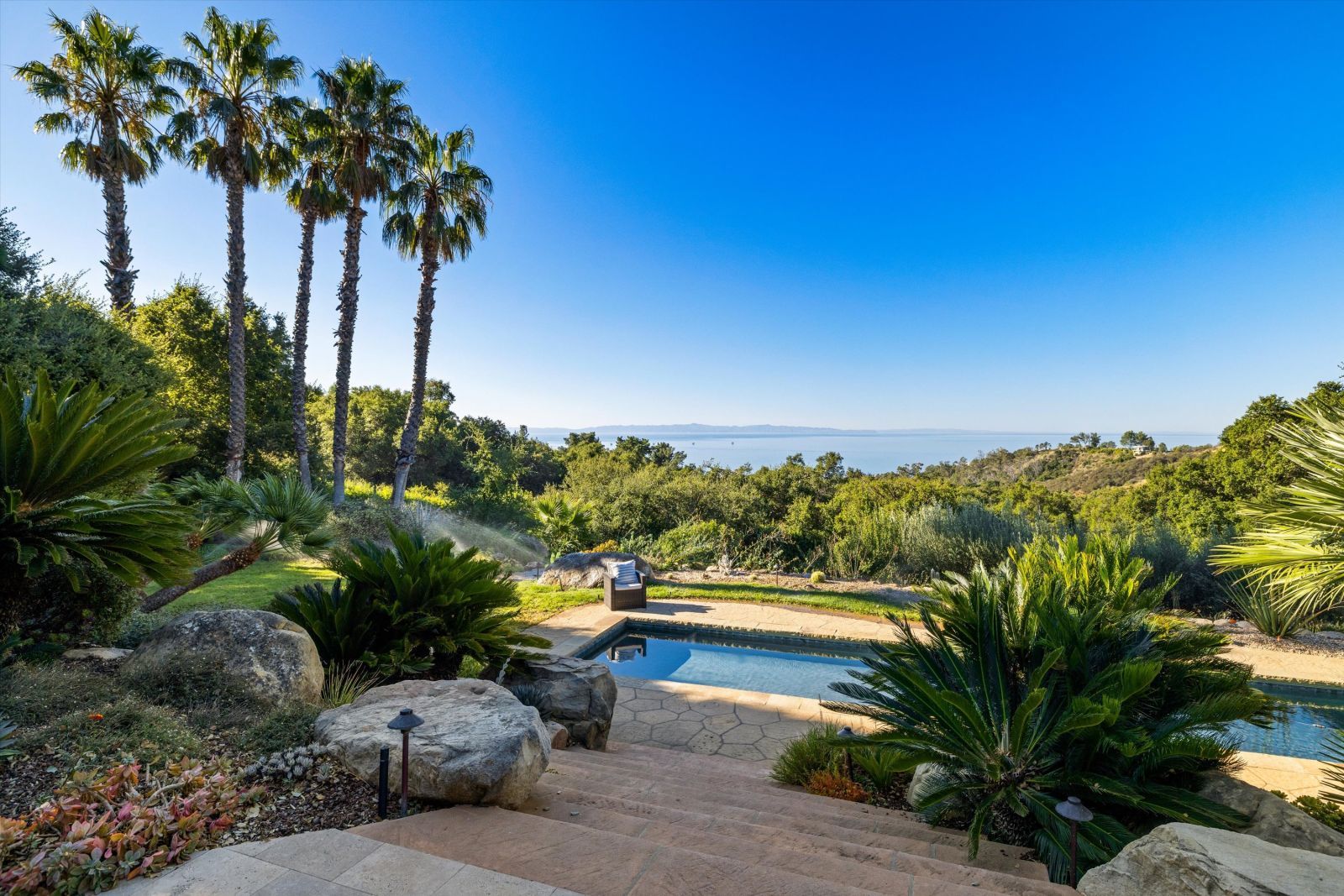 Palm trees stand tall in this lushly landscaped backyard, with a sparkling pool, expansive terrace, palm trees. and ocean on the horizon.